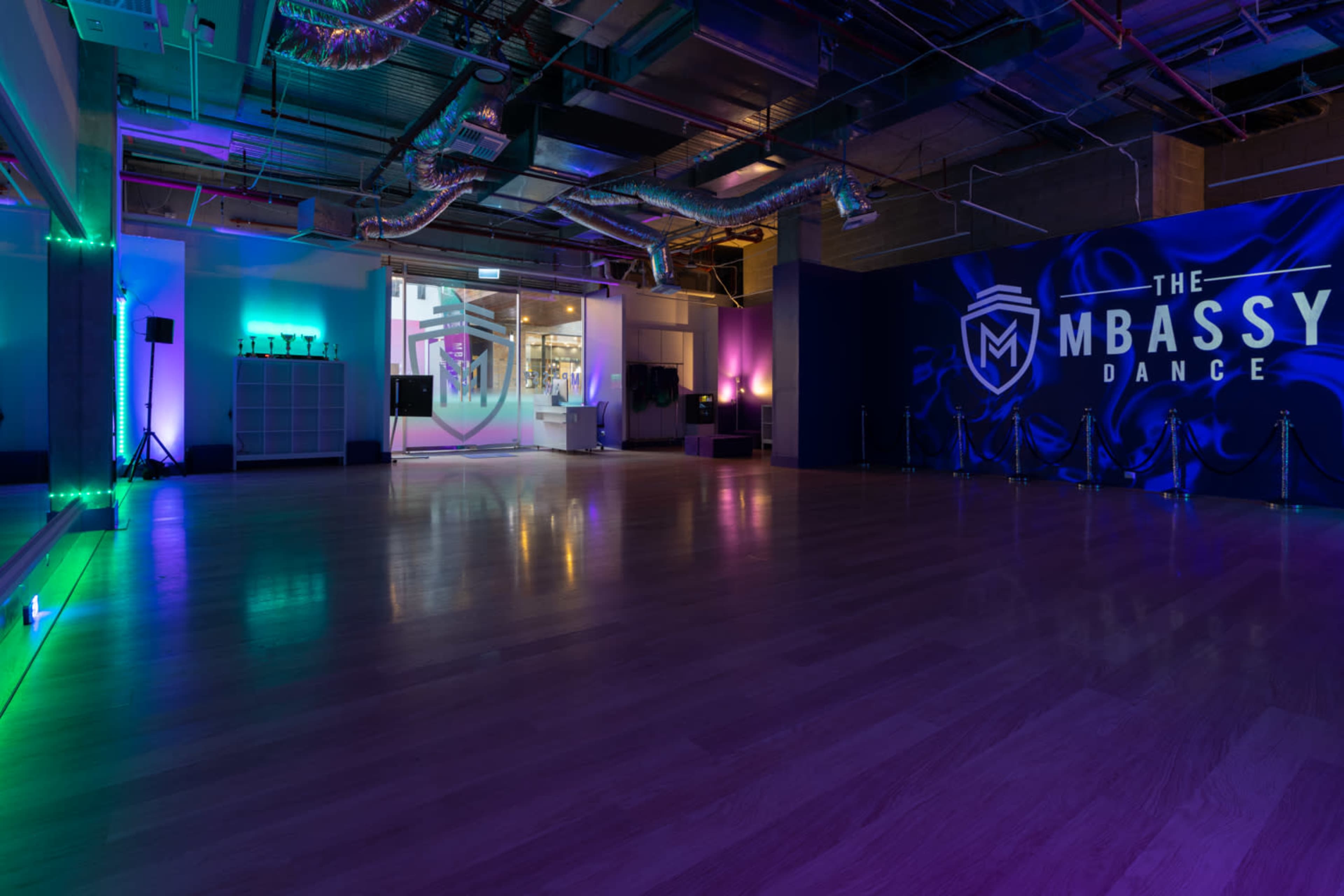 A spacious dance studio with a wooden floor, illuminated by colorful lights and featuring a large wall logo that reads "THE MBASSY DANCE."