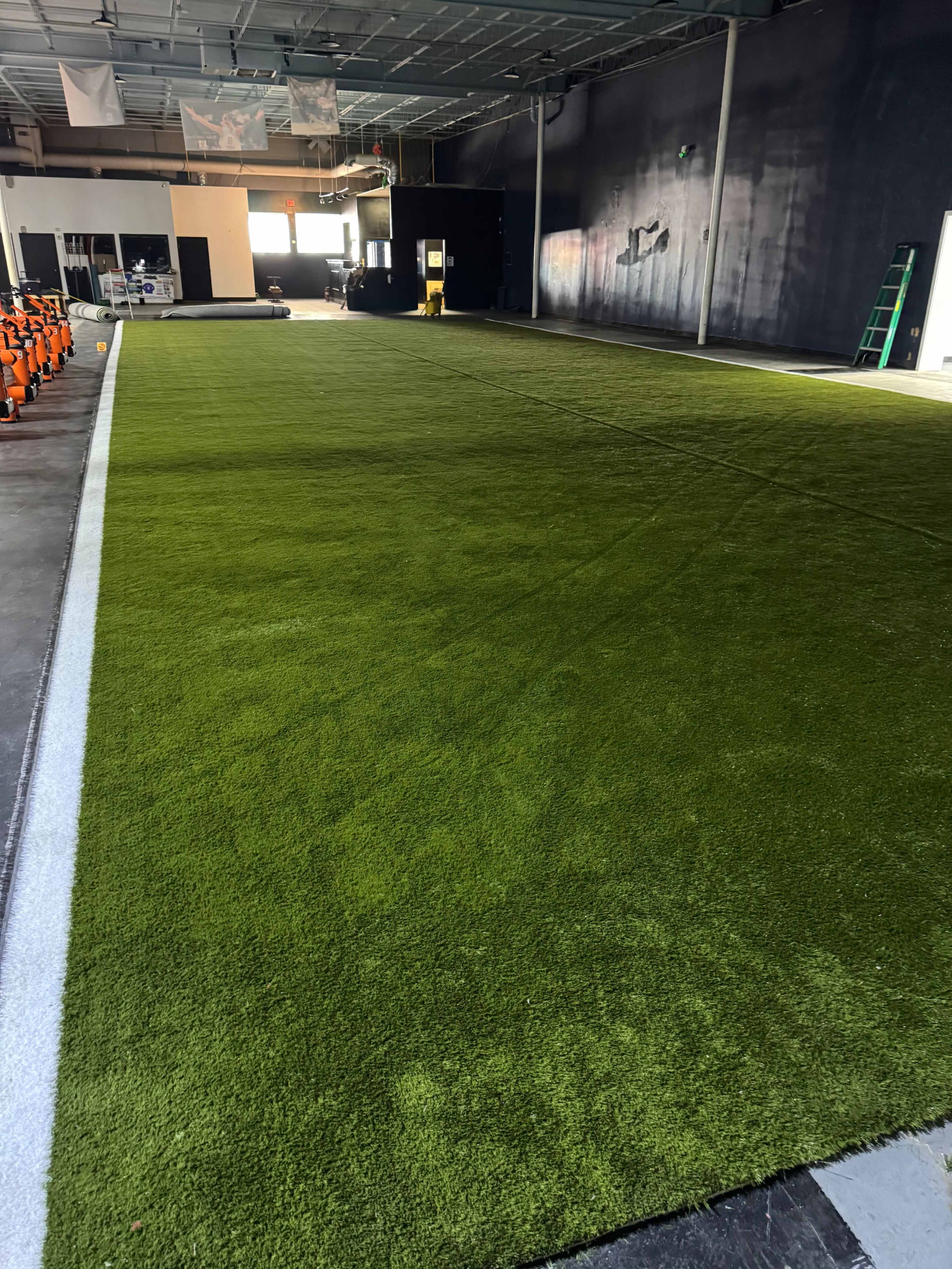 The image shows a large indoor space with newly installed green artificial turf, surrounded by a dark wall and equipment in the background.