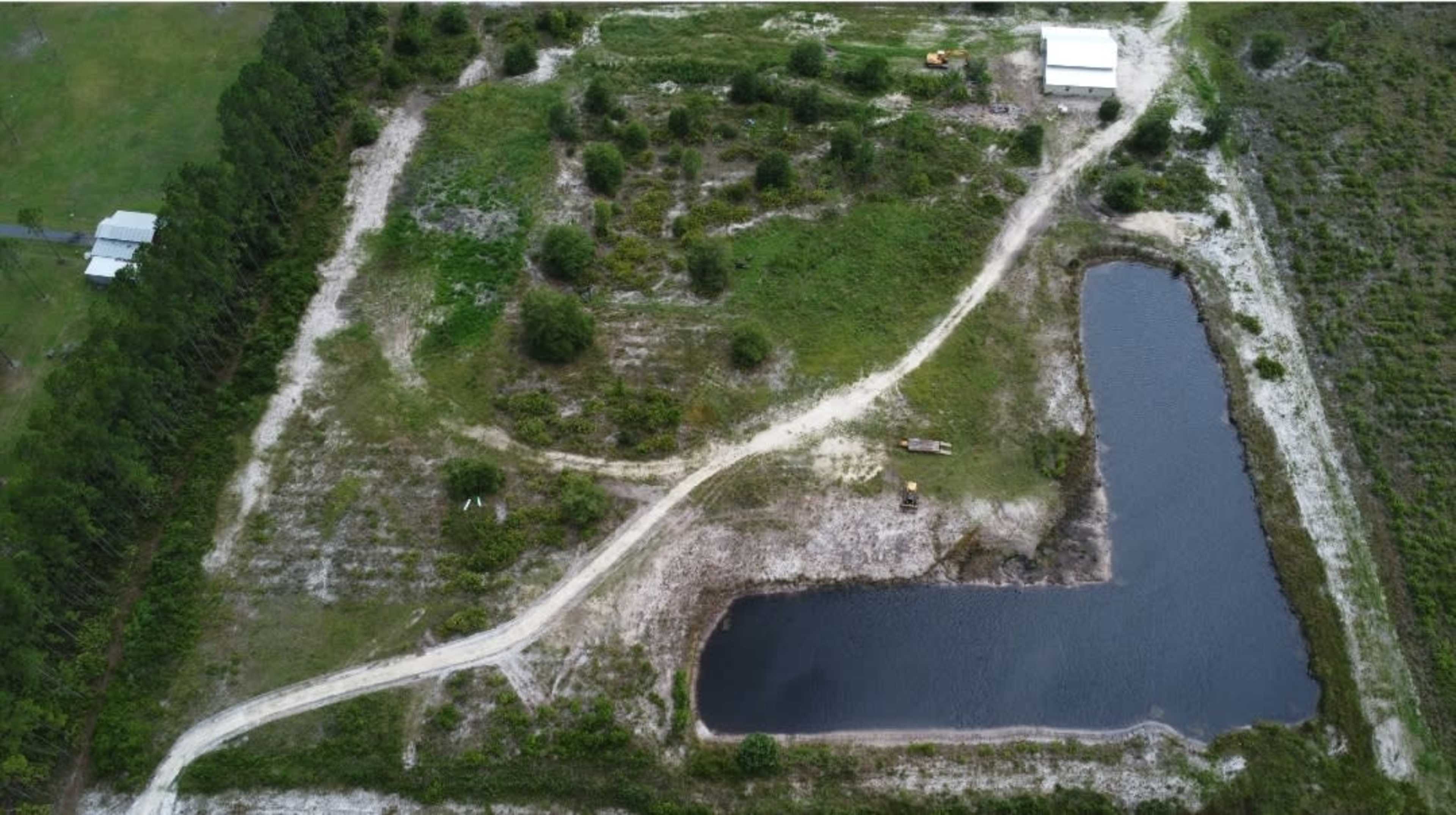 An aerial view of a large, vacant plot of land with a winding dirt path, a small pond, and some scattered trees.