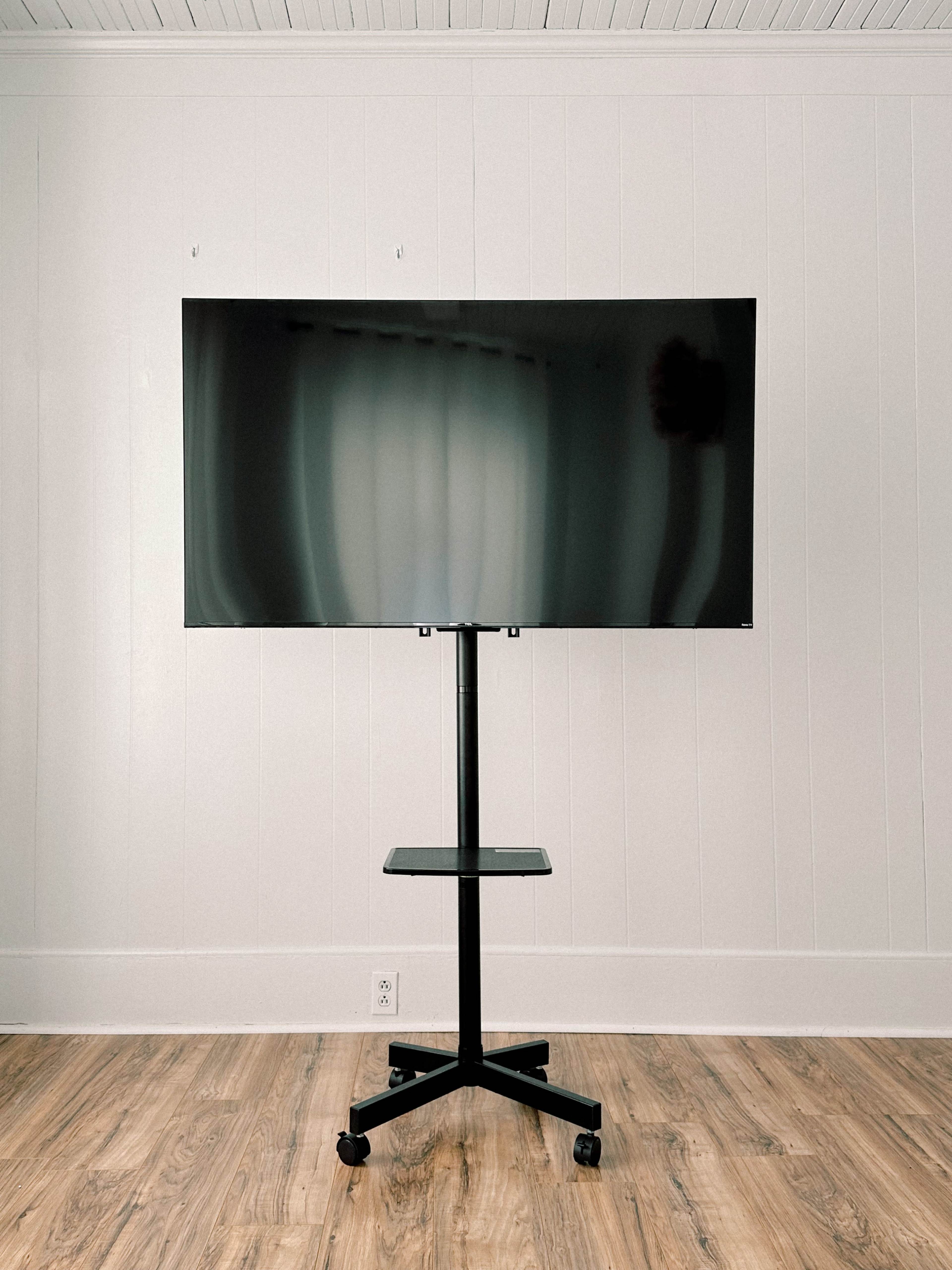 A large curved monitor is mounted on a black rolling stand in a room with wooden flooring and white walls.