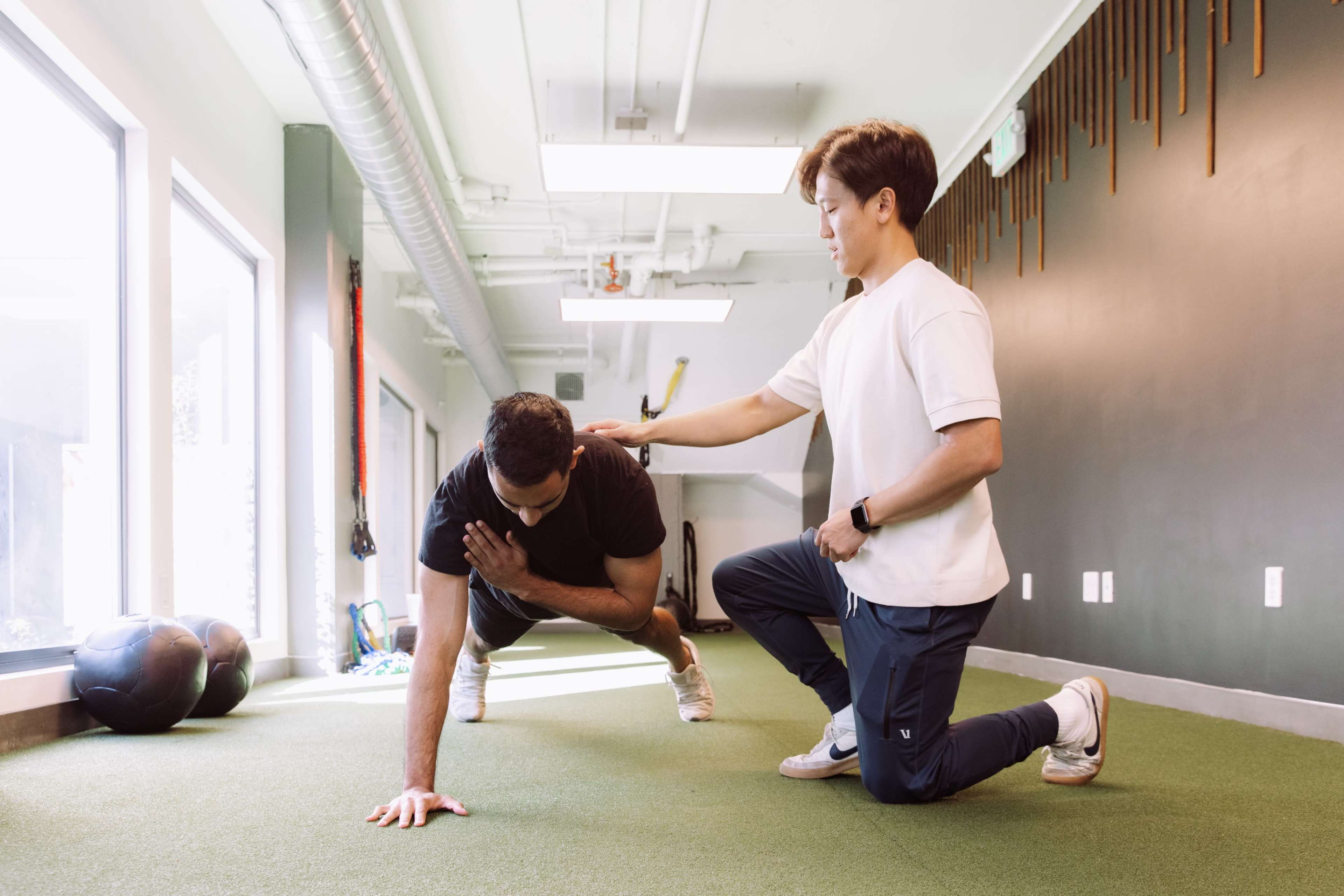 A trainer assists a person performing a push-up on a green gym floor.