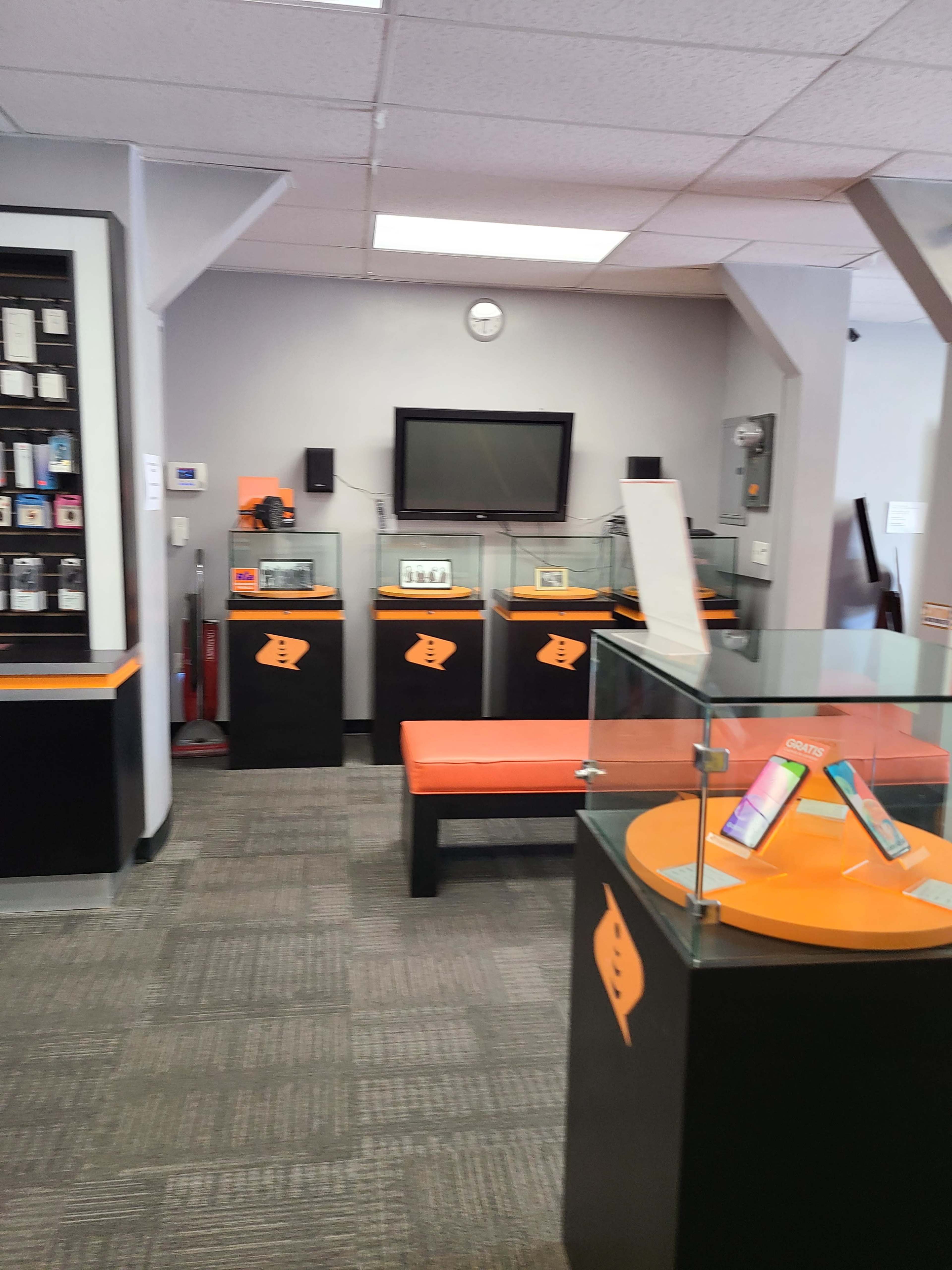 A retail store interior featuring display cases with electronic devices, a bench, a wall-mounted TV, and a clock.