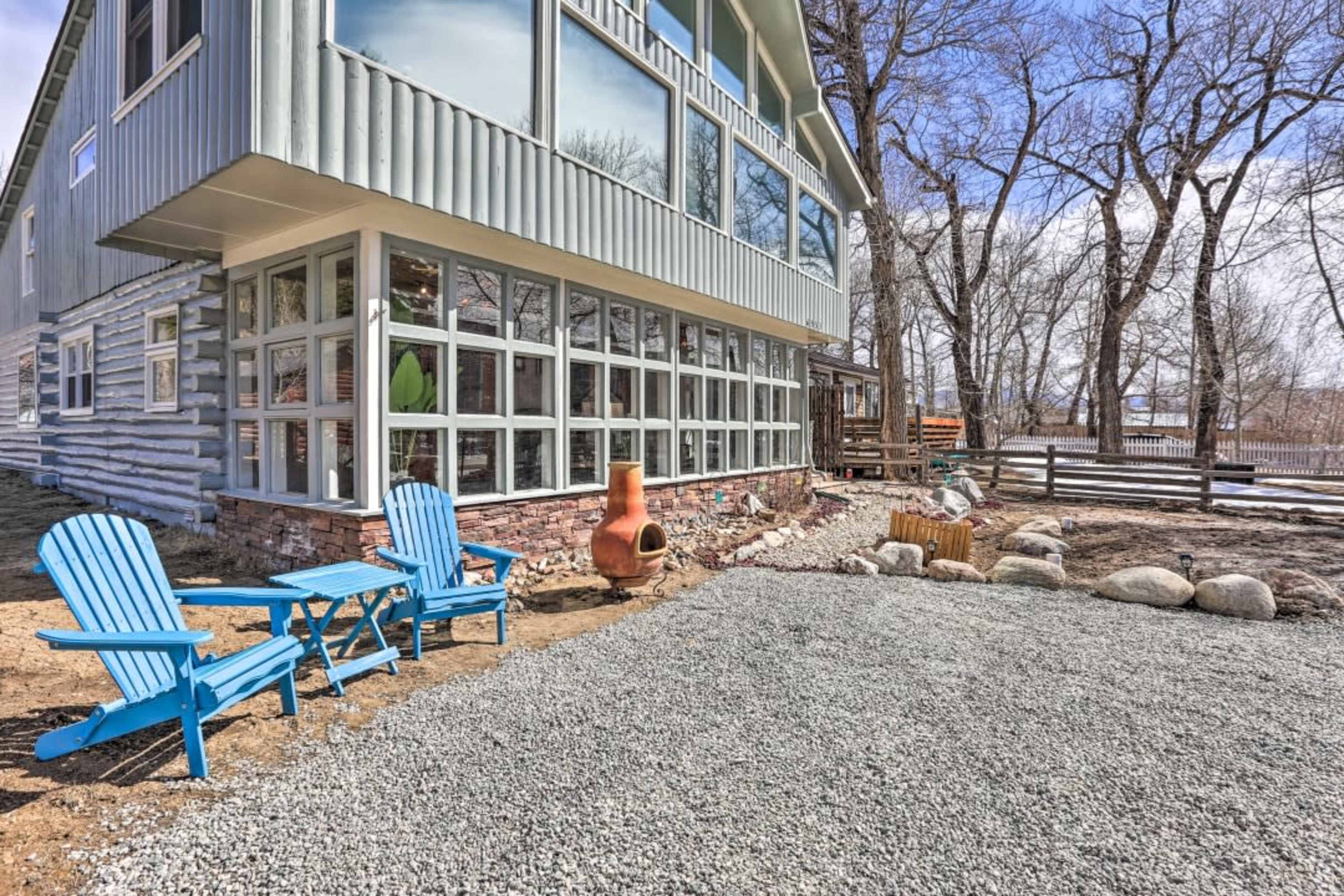 A home with a large glass wall features two blue adirondack chairs and a gravel area near a chimney in a yard surrounded by trees.