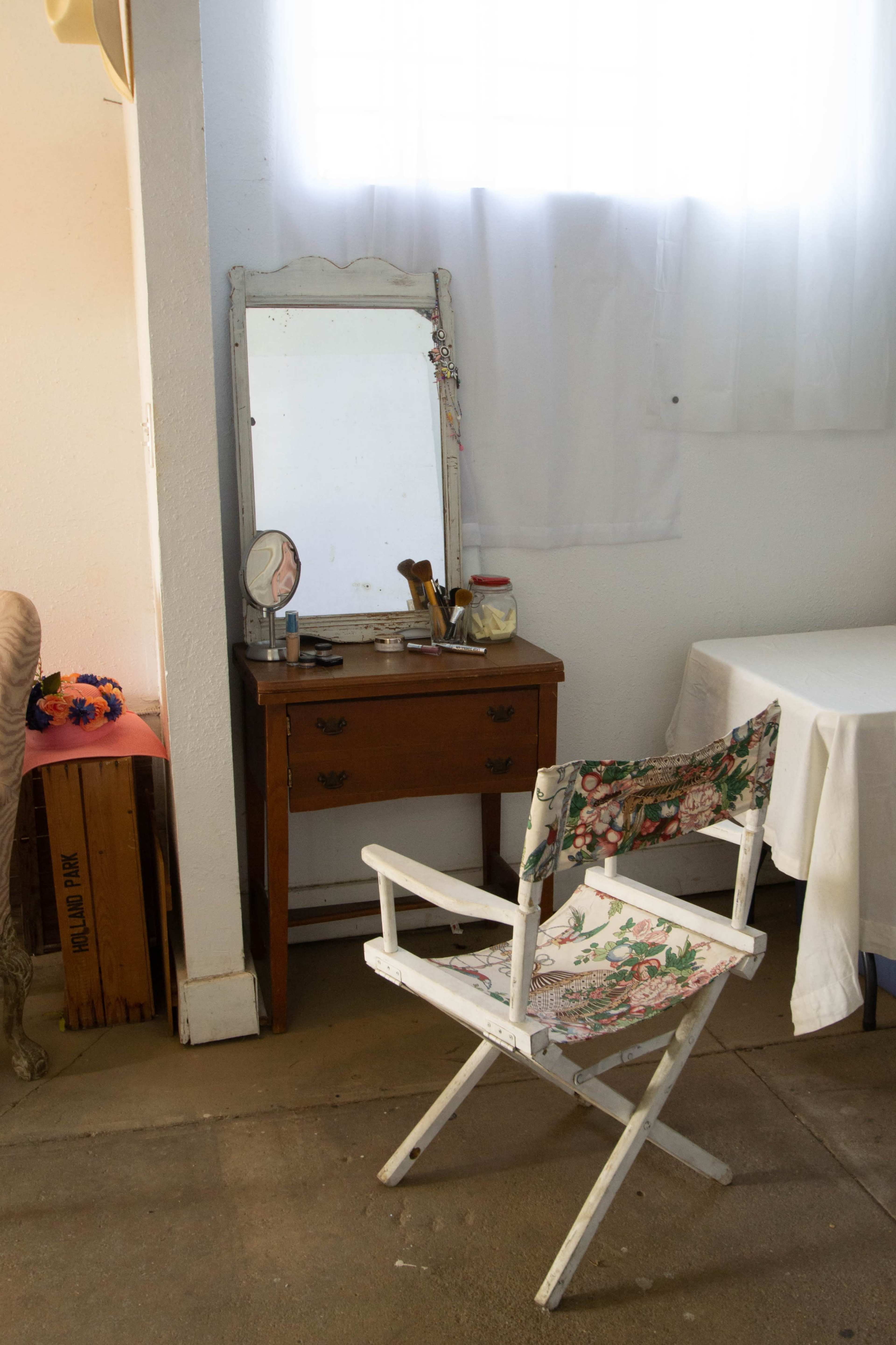 A wooden dresser with a mirror sits next to a chair featuring a floral fabric, in a room with white curtains and a table.