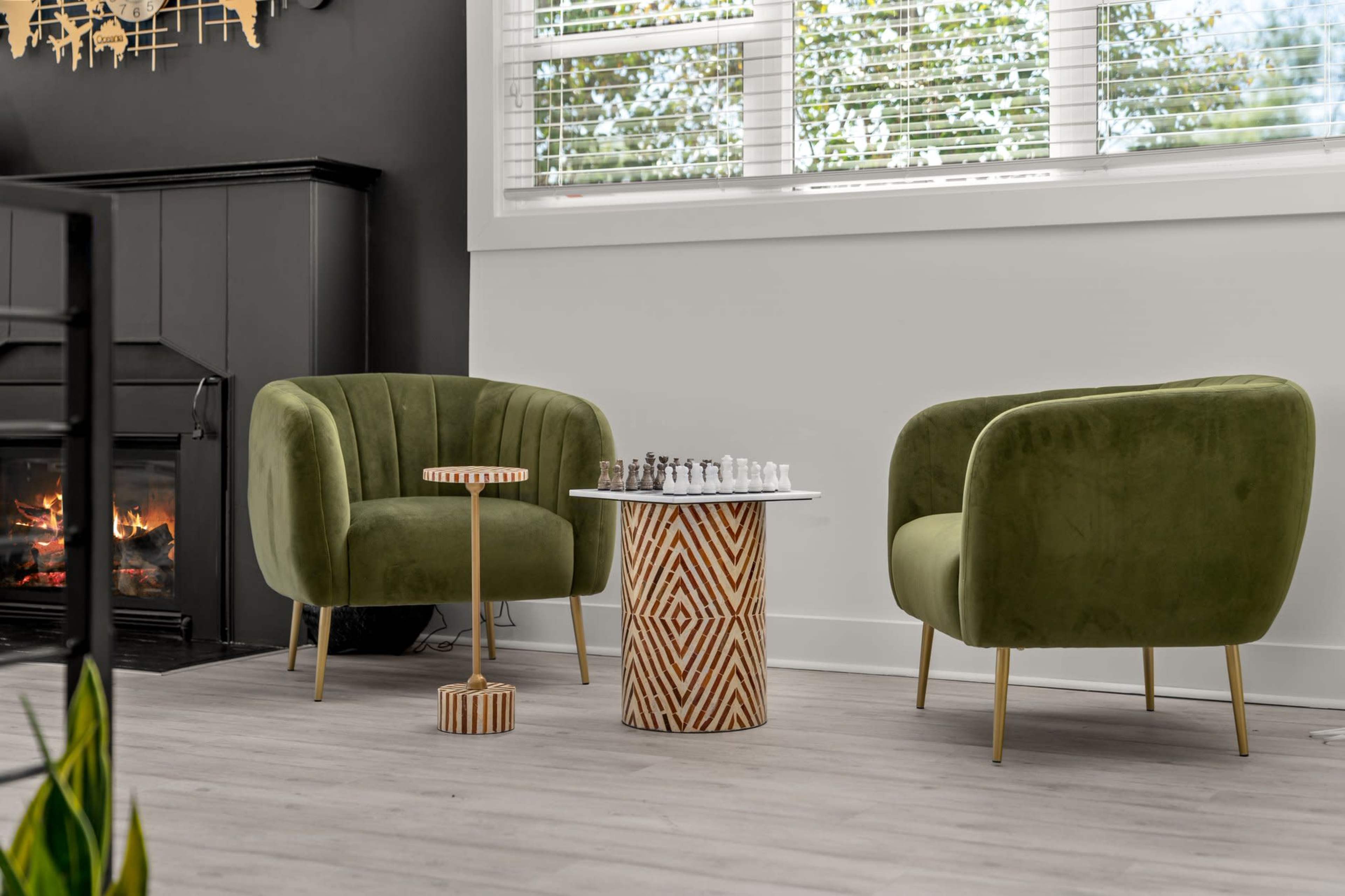 The image shows a cozy living space featuring two green upholstered chairs, a round patterned table with a chess set, and a fireplace in the background.