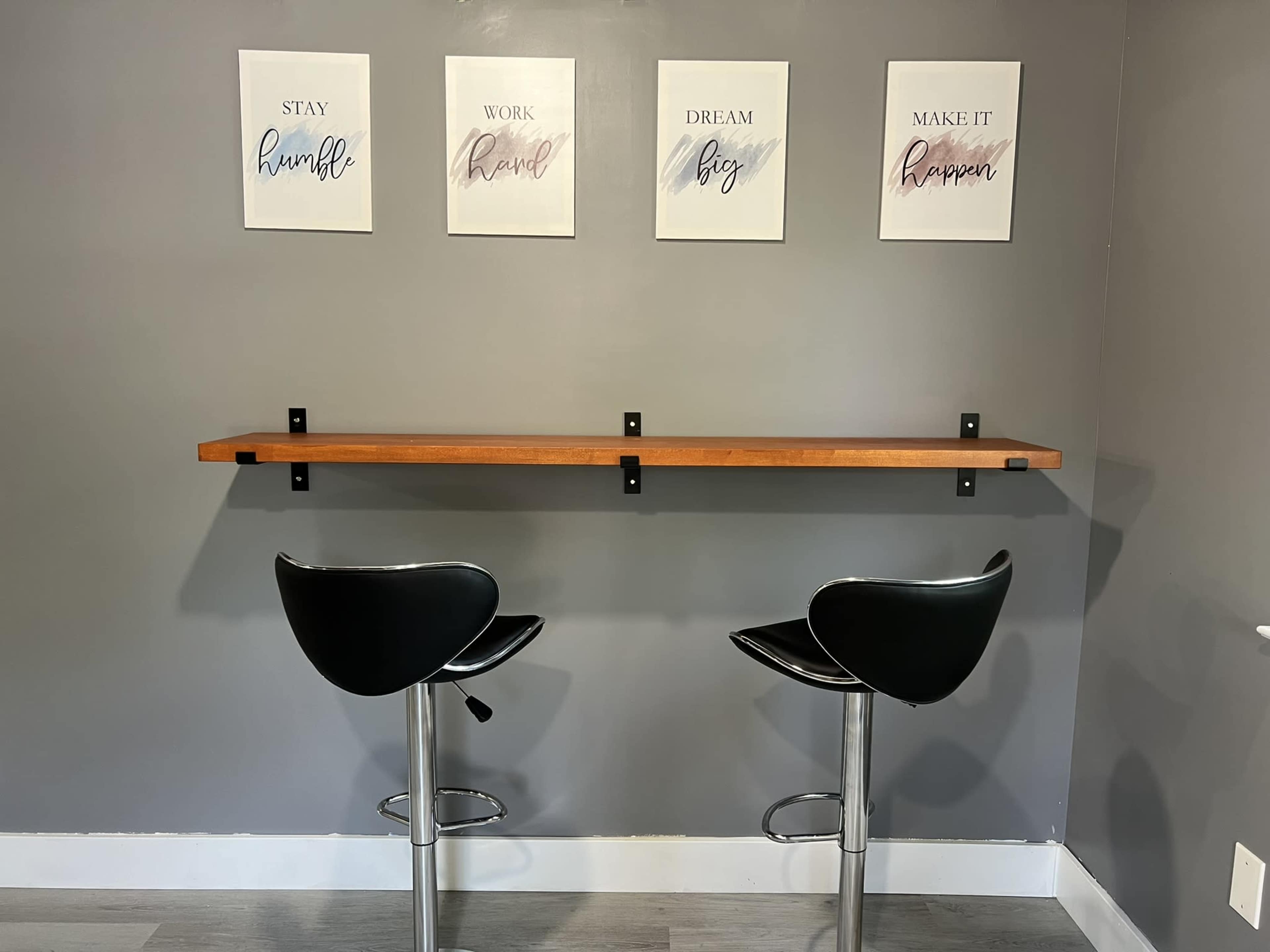 A minimalist workspace features a narrow wooden shelf mounted on a gray wall, with two black bar stools positioned underneath and four framed motivational prints above.