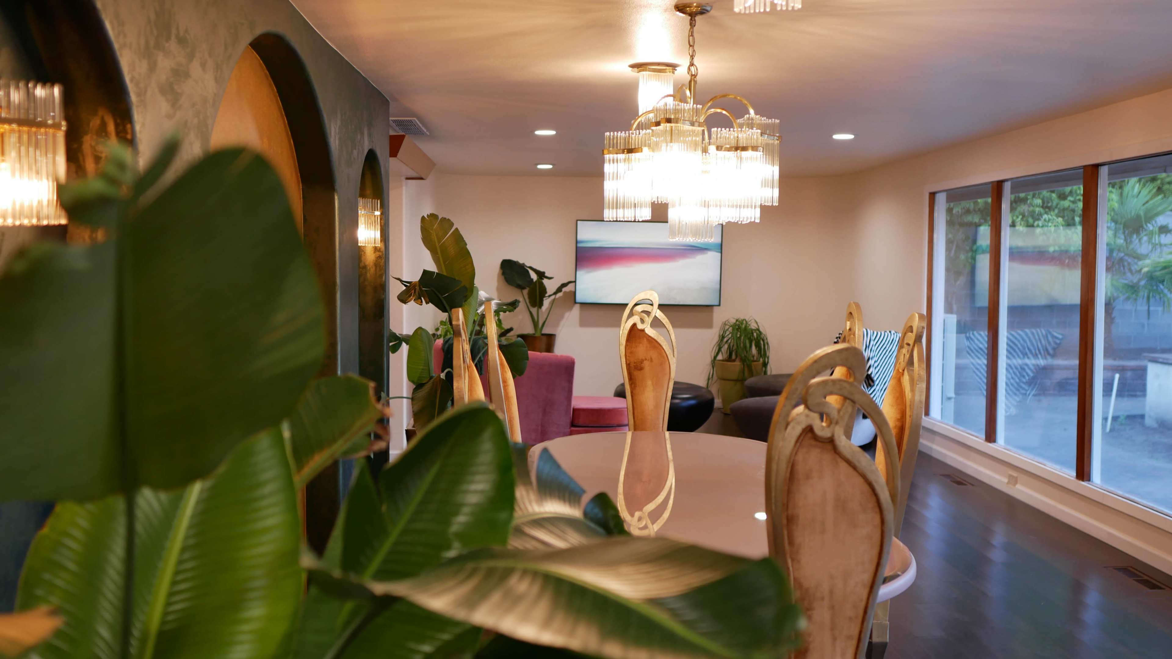 A dining area with a large table surrounded by ornate chairs, illuminated by a chandelier, and flanked by plants and a modern artwork in the background.