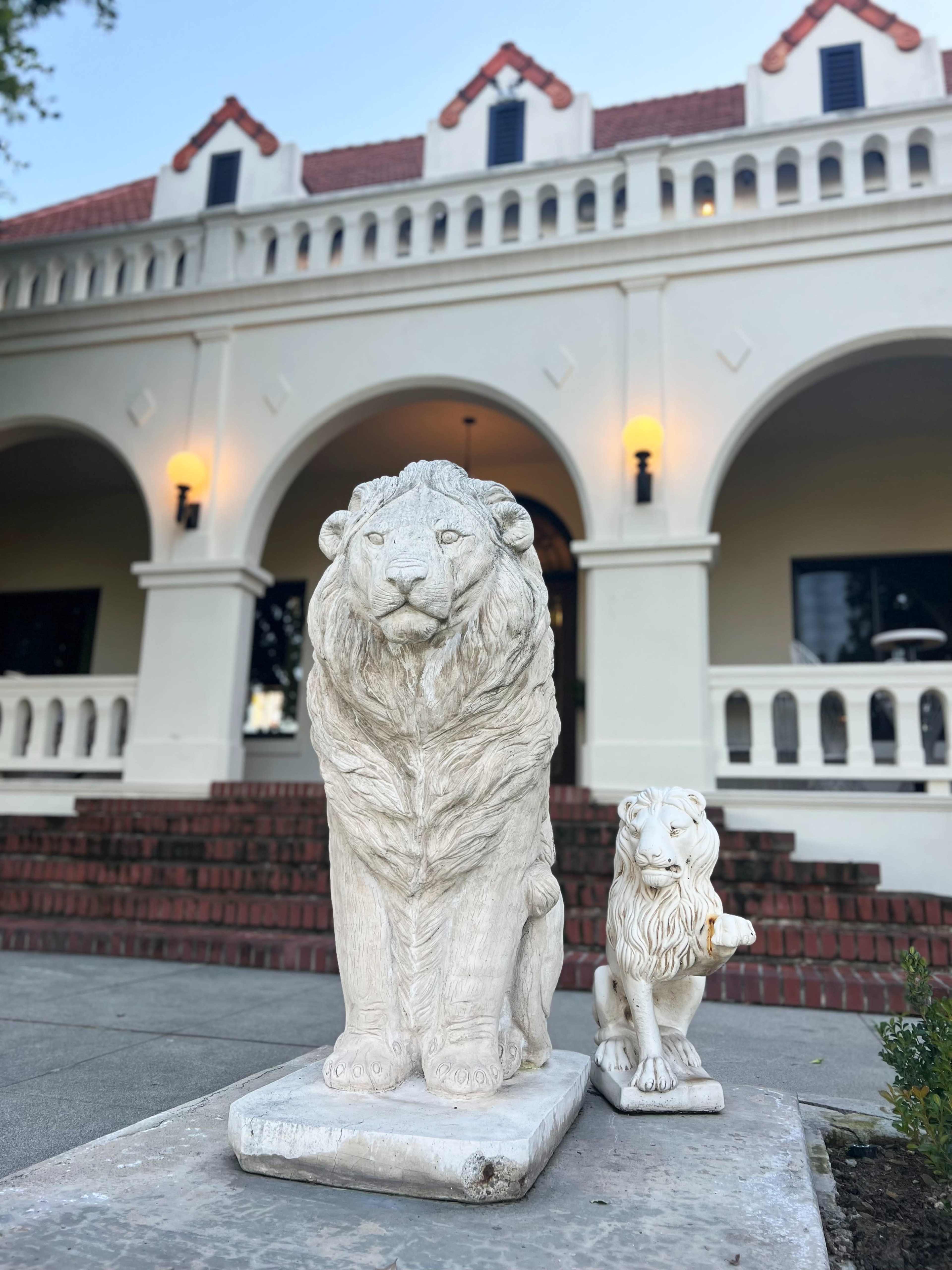 Two lion statues, one large and one small, stand in front of a white building with arched windows and red brick steps.