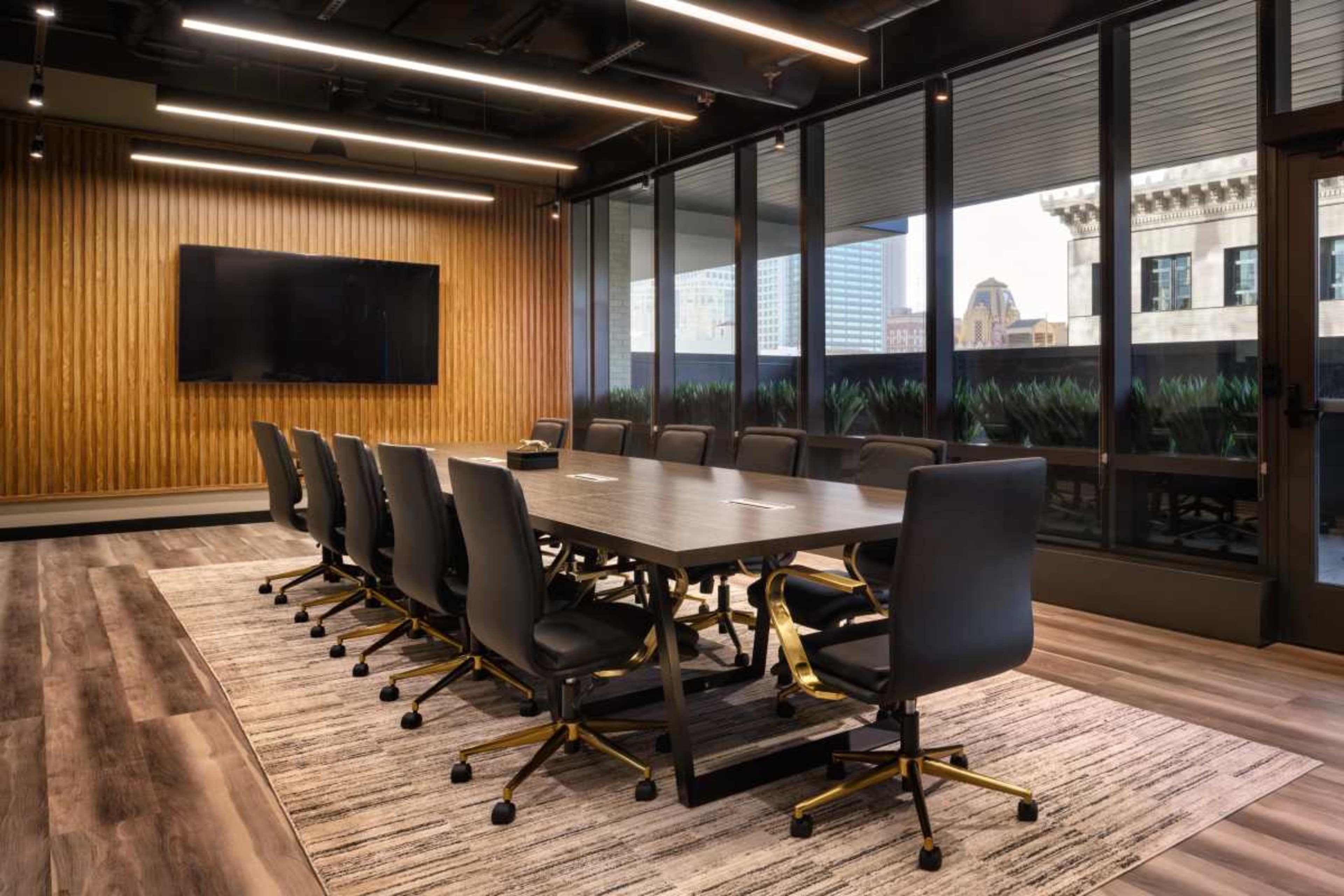 A modern conference room features a large table surrounded by black chairs, centered under overhead lighting and framed by large windows.