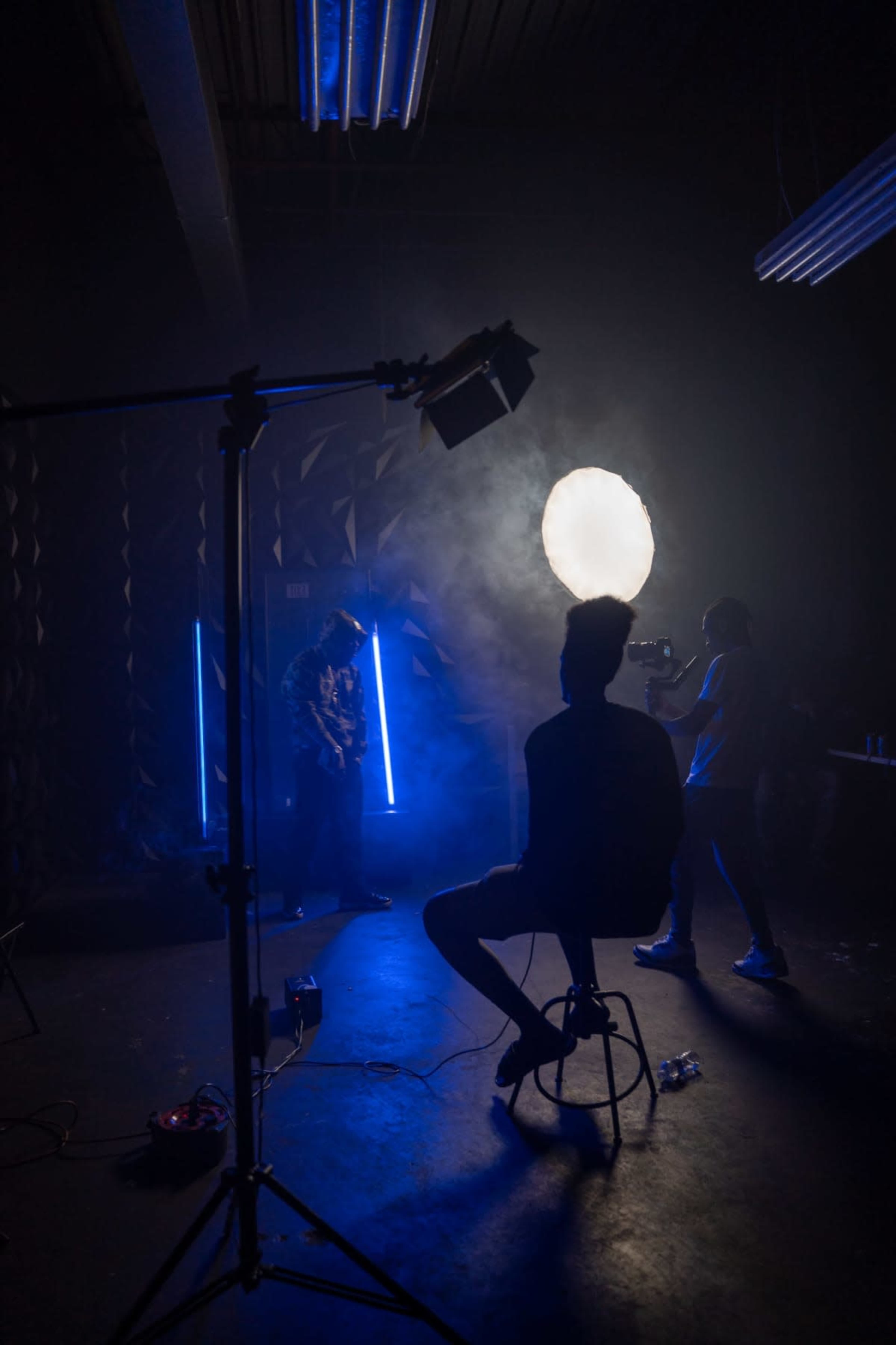 A silhouetted person sits on a stool while another person photographs a musician illuminated by bright lights in a dimly lit studio.