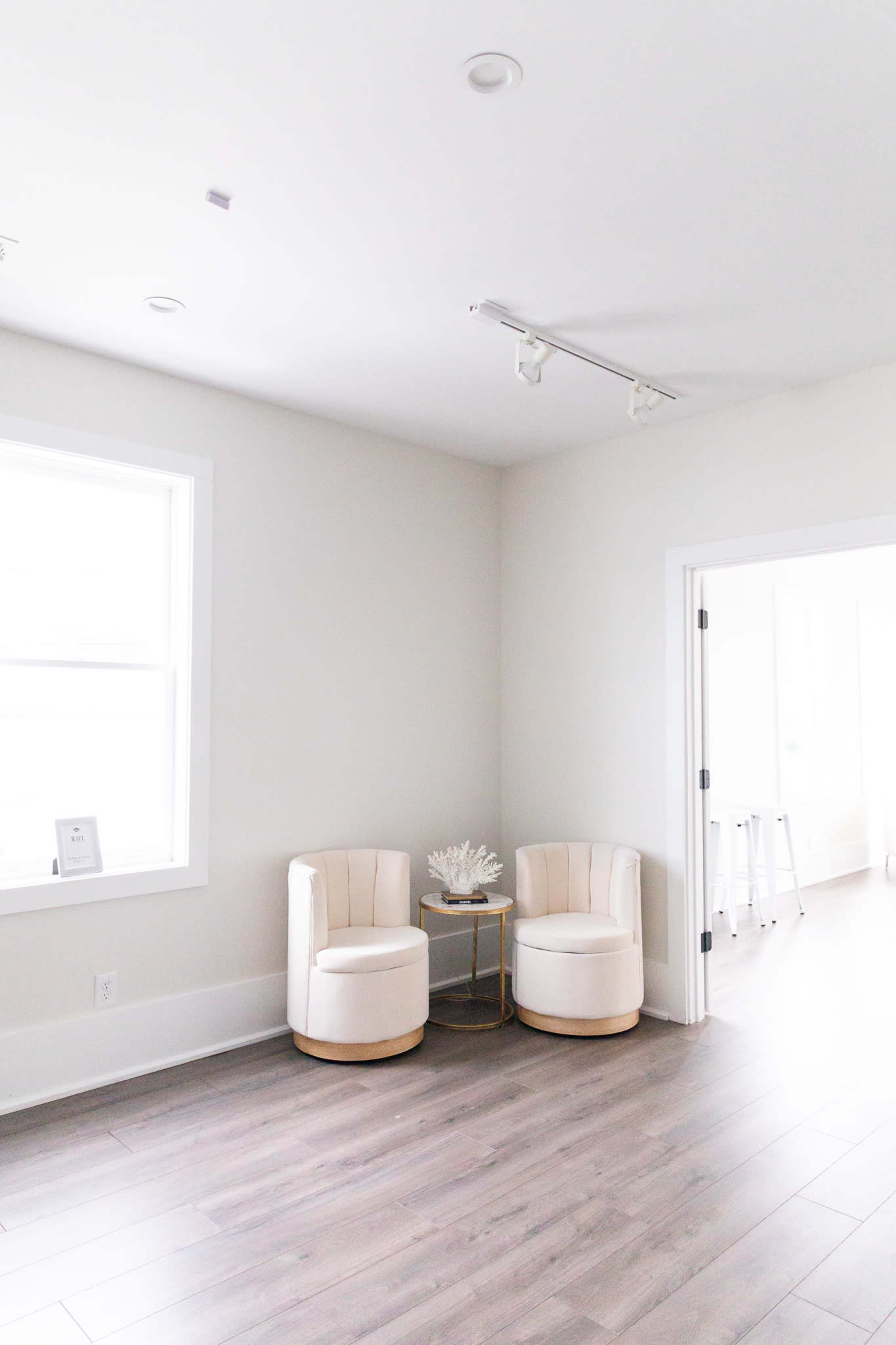 A bright, minimalistic room features two cream-colored chairs and a small side table between them, with light wood flooring and a window letting in natural light.