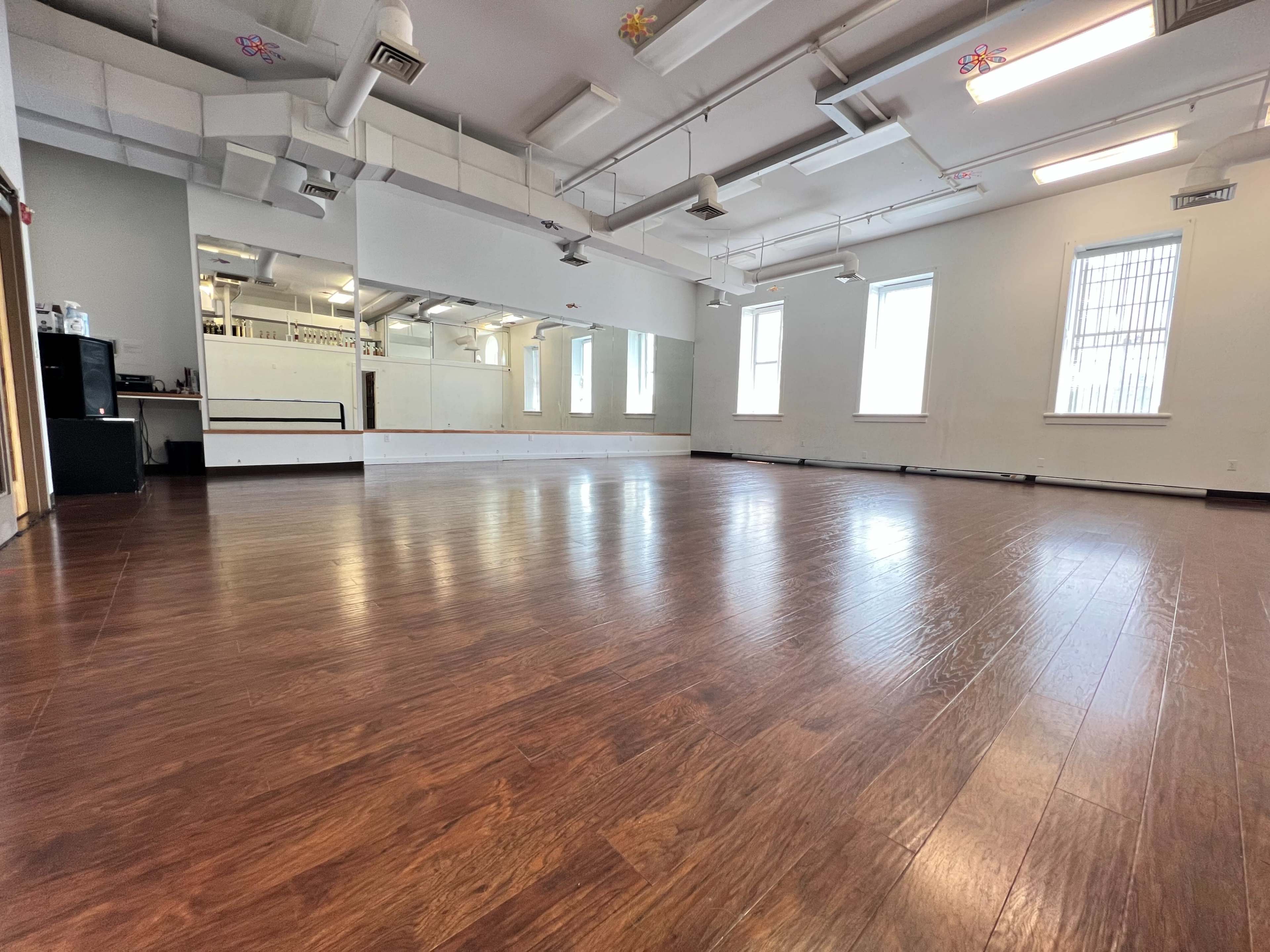 An empty dance studio with wooden flooring, large mirrors, and multiple windows allowing natural light to enter.