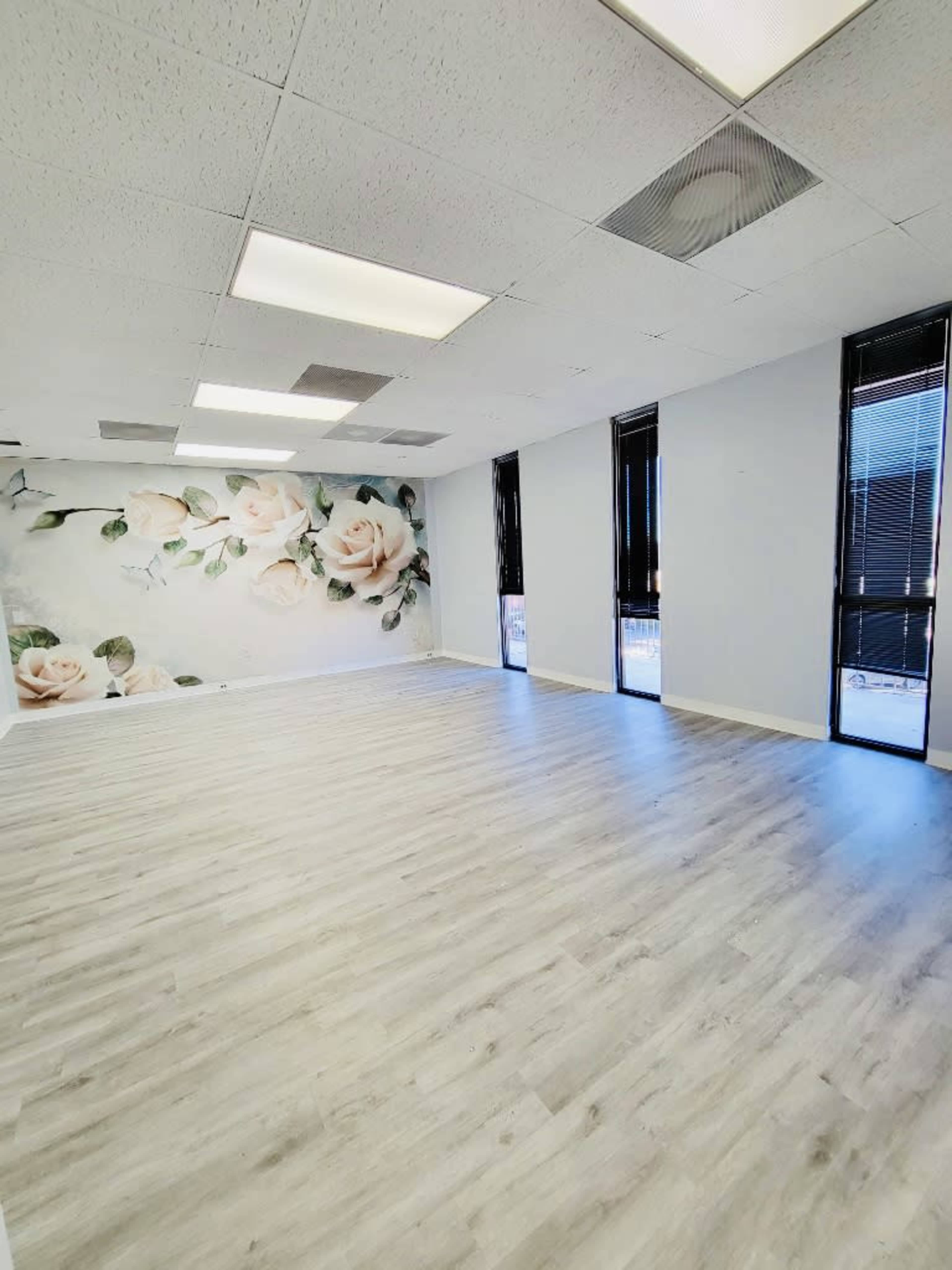 A spacious, empty room with light wooden flooring, large windows, and a wall featuring a floral mural.