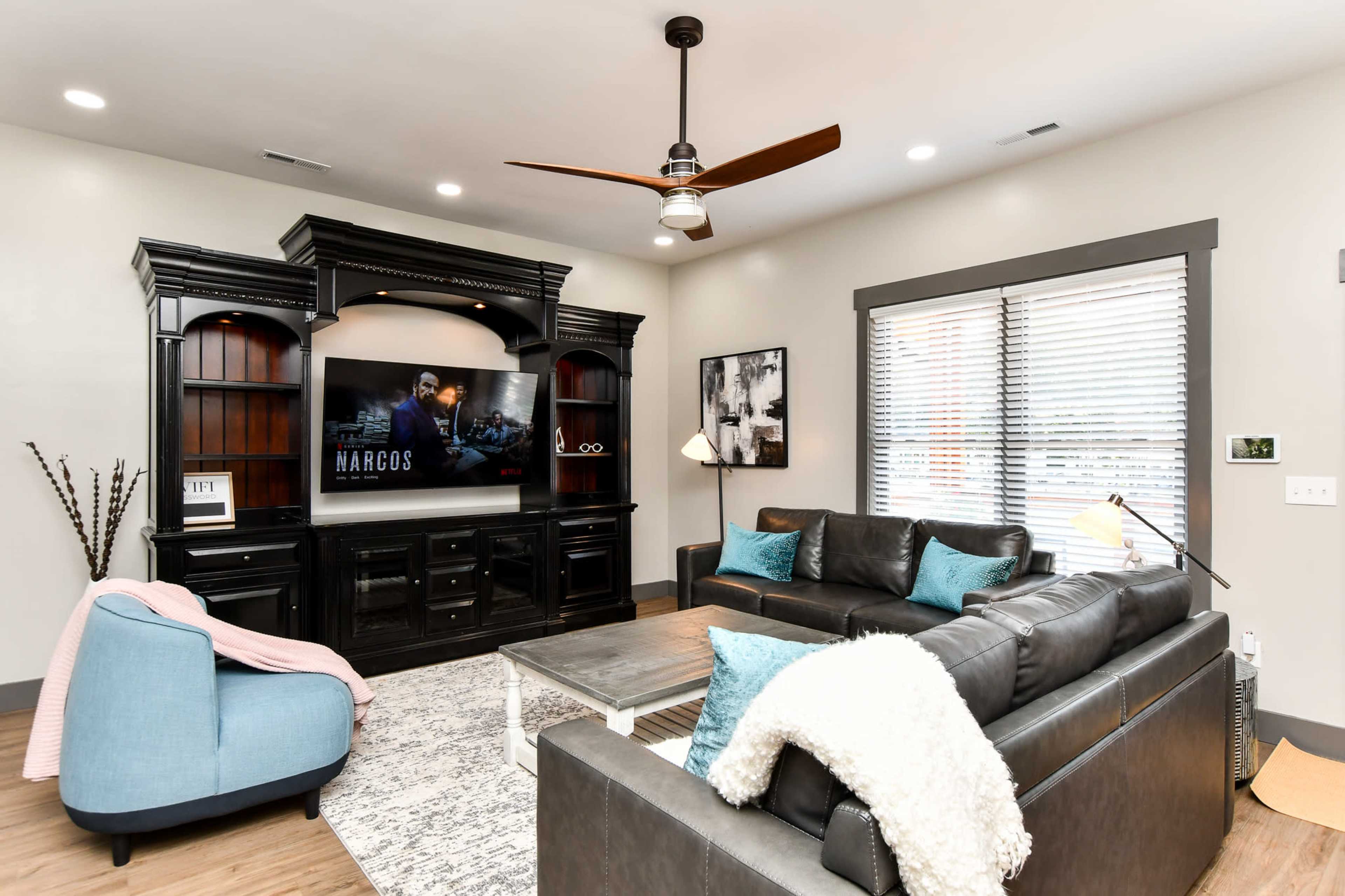 The living room features a dark wooden entertainment center with a mounted TV displaying a show, a black leather sofa with decorative pillows, and a light-colored area rug.