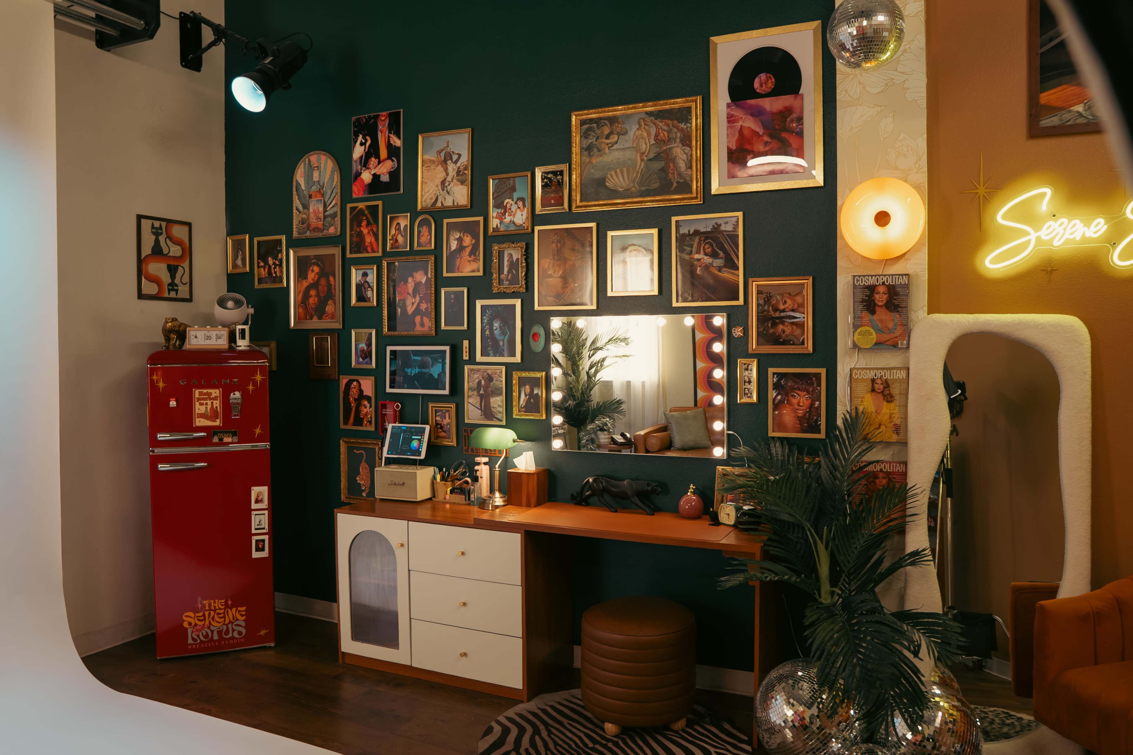 The image features a stylish studio space with a green wall adorned with various framed photos, a wooden vanity with a large mirror, a vintage red refrigerator, and decorative lighting.
