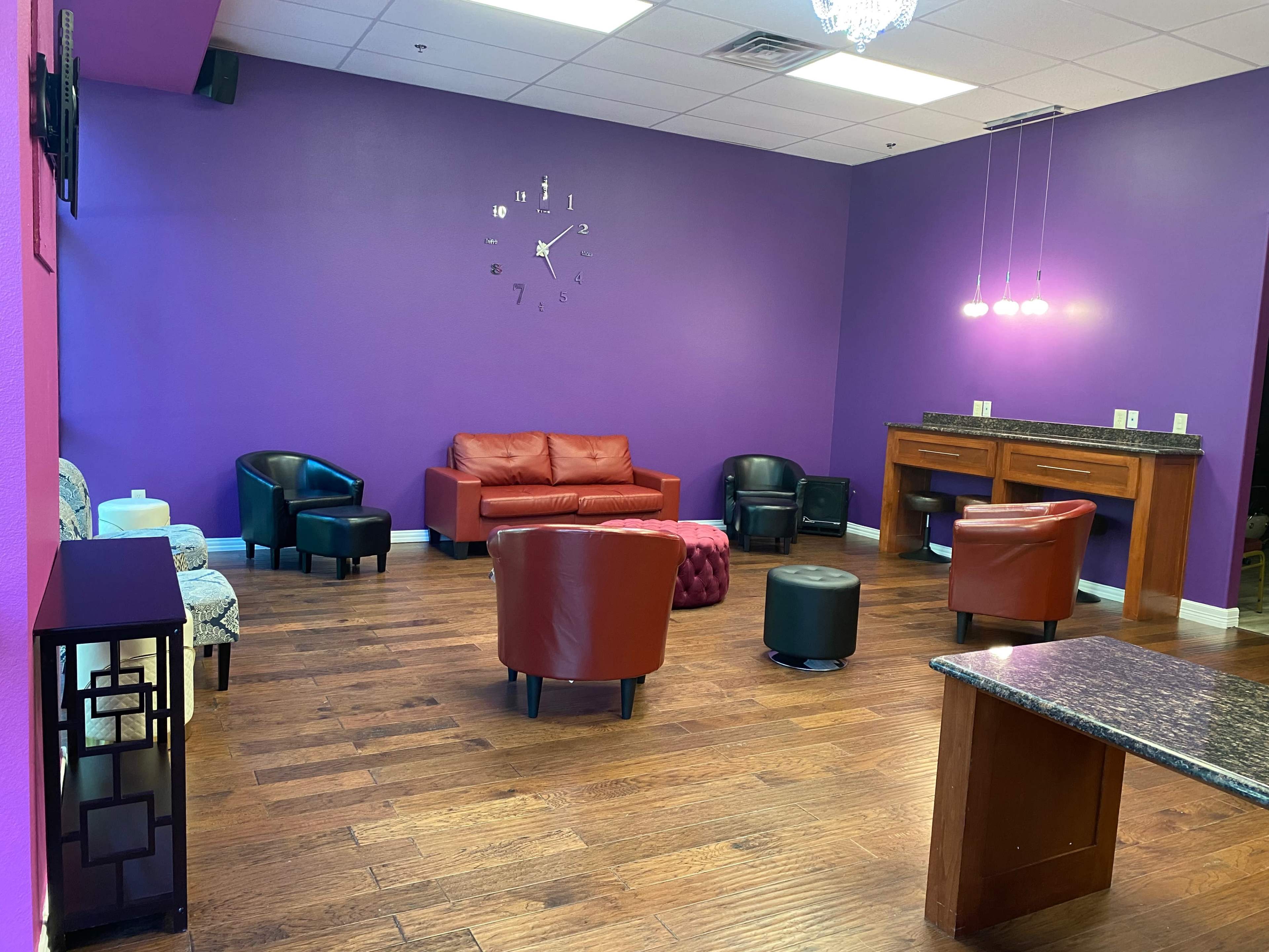 A lounge area with a mix of seating options, including a red sofa, several black chairs, and a few small tables, all set against a vibrant purple wall and wooden flooring.