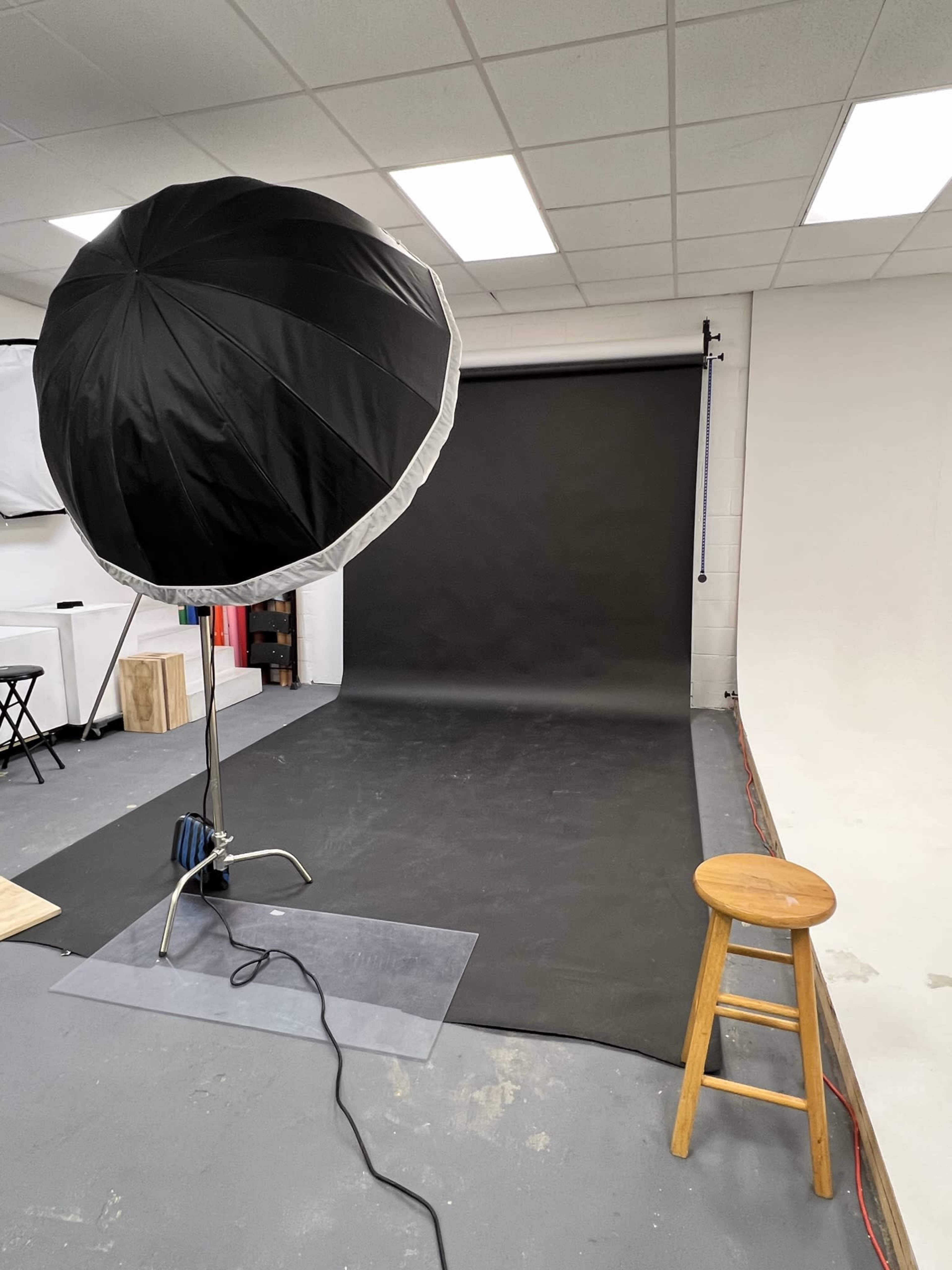 A photography studio with a large softbox light on a stand, a black backdrop, and a wooden stool positioned on a gray floor.