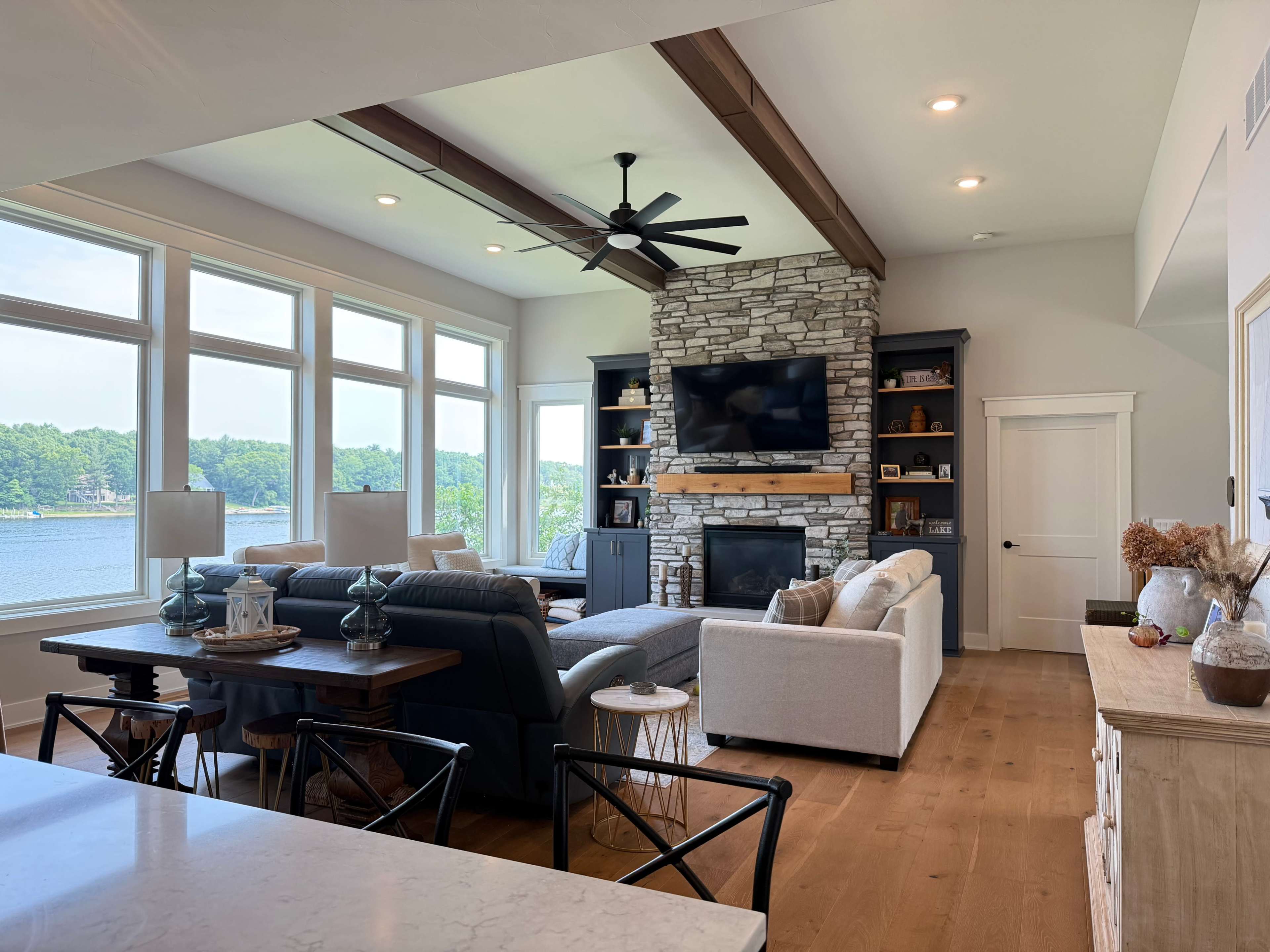 The image shows a modern living room with large windows overlooking a lake, featuring a stone fireplace, comfortable seating, and a dining area.