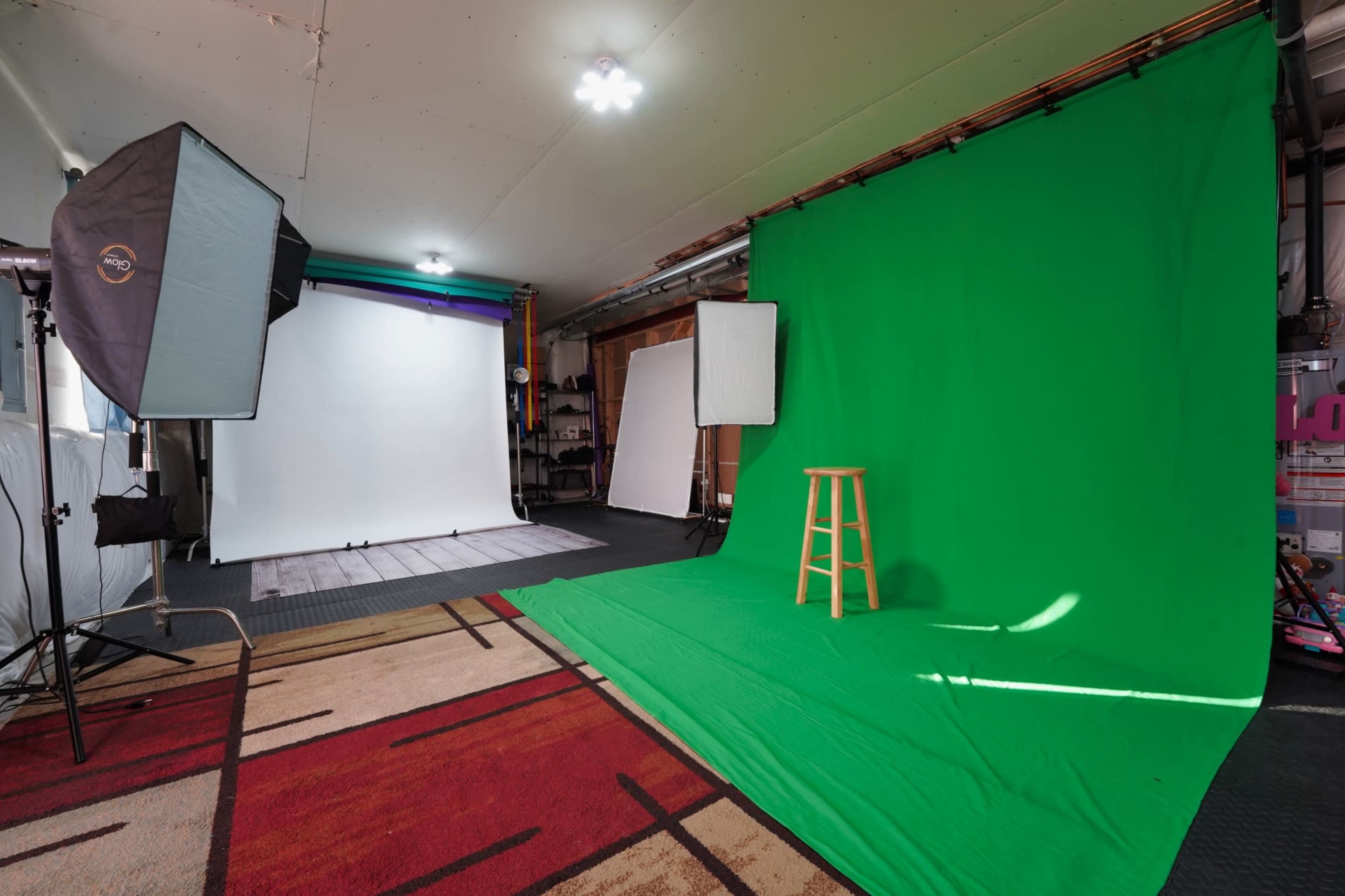The image shows a photography studio featuring two backdrops: one green and one white, along with softbox lights and a wooden stool positioned in front of the green backdrop.
