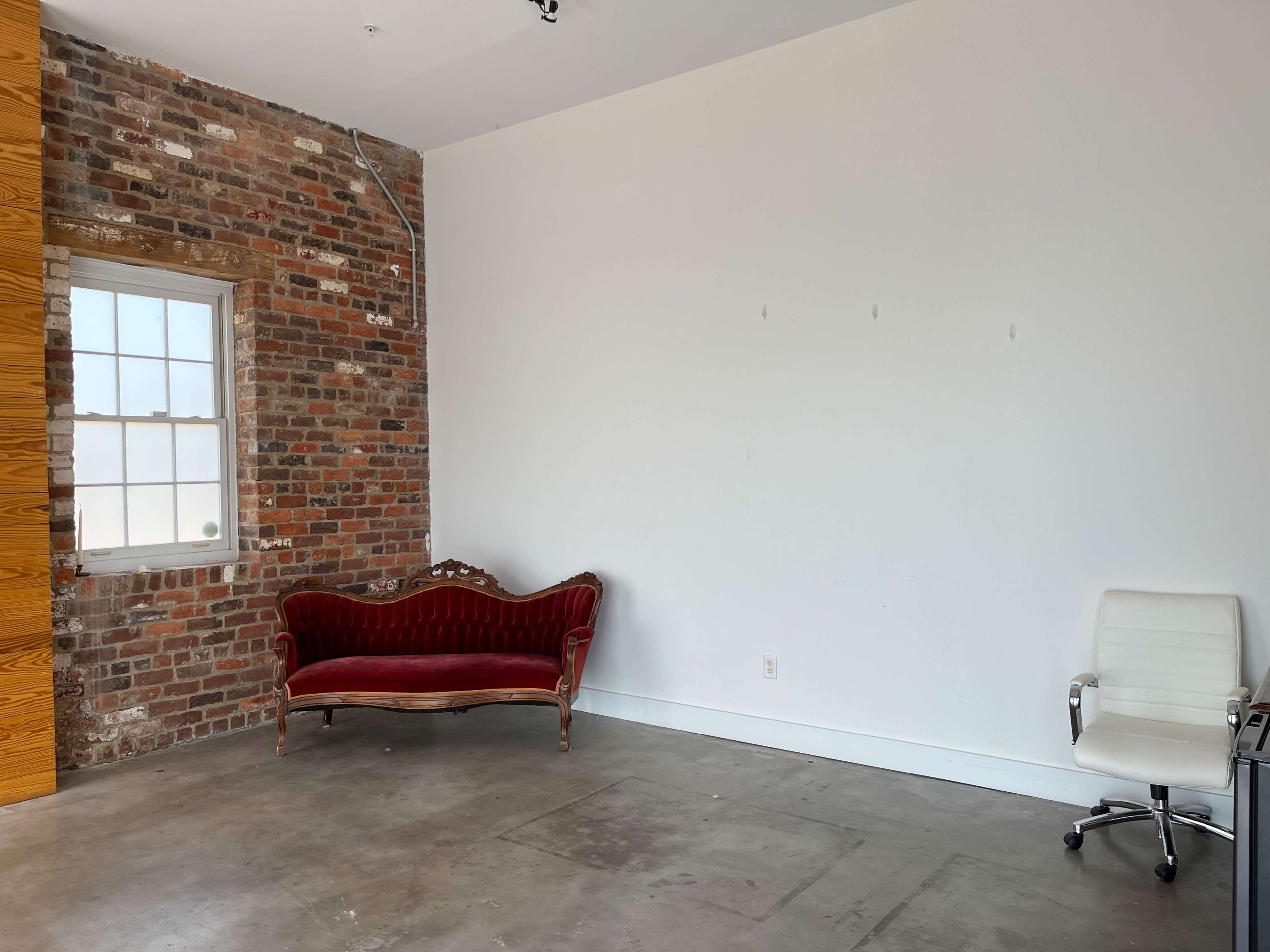 The image shows a room with a brick wall on one side, featuring a red velvet sofa and a white office chair on a concrete floor.