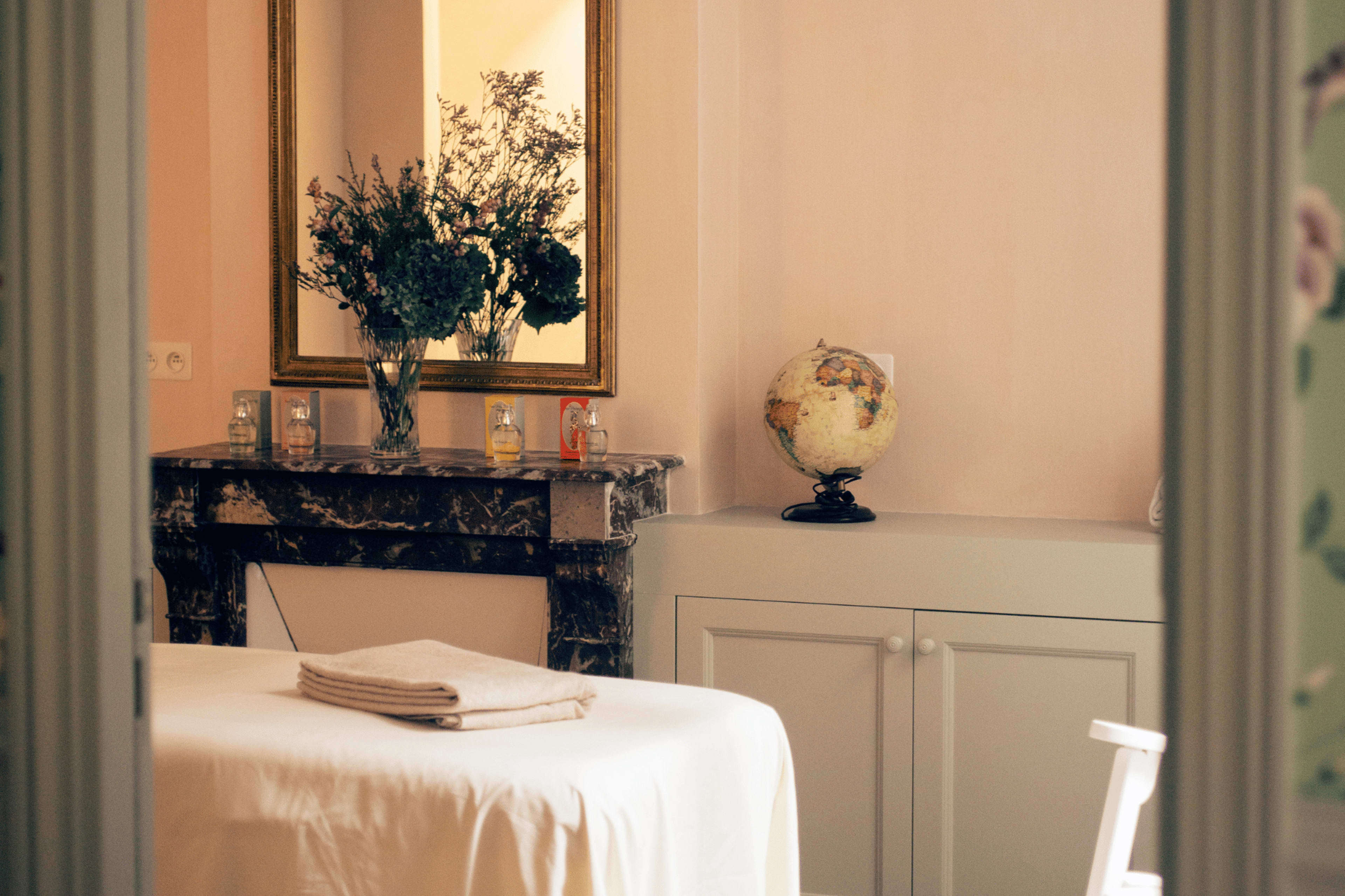 A massage table covered with a folded towel is positioned in a softly lit room that features a mirror and a globe on a marble mantelpiece.