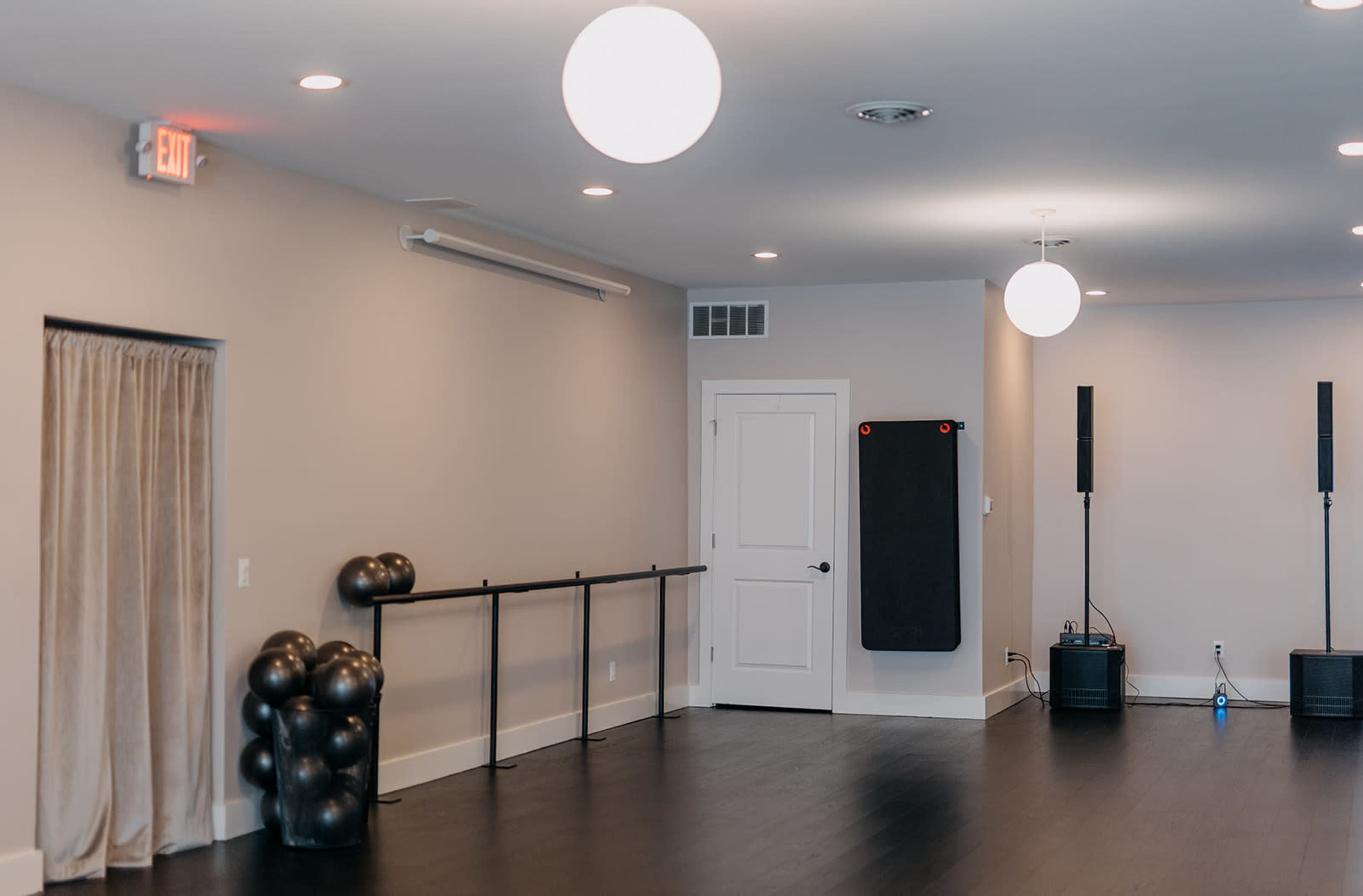 A spacious room with a wooden floor, featuring a ballet bar on one wall, a door, and black exercise balls in the corner.