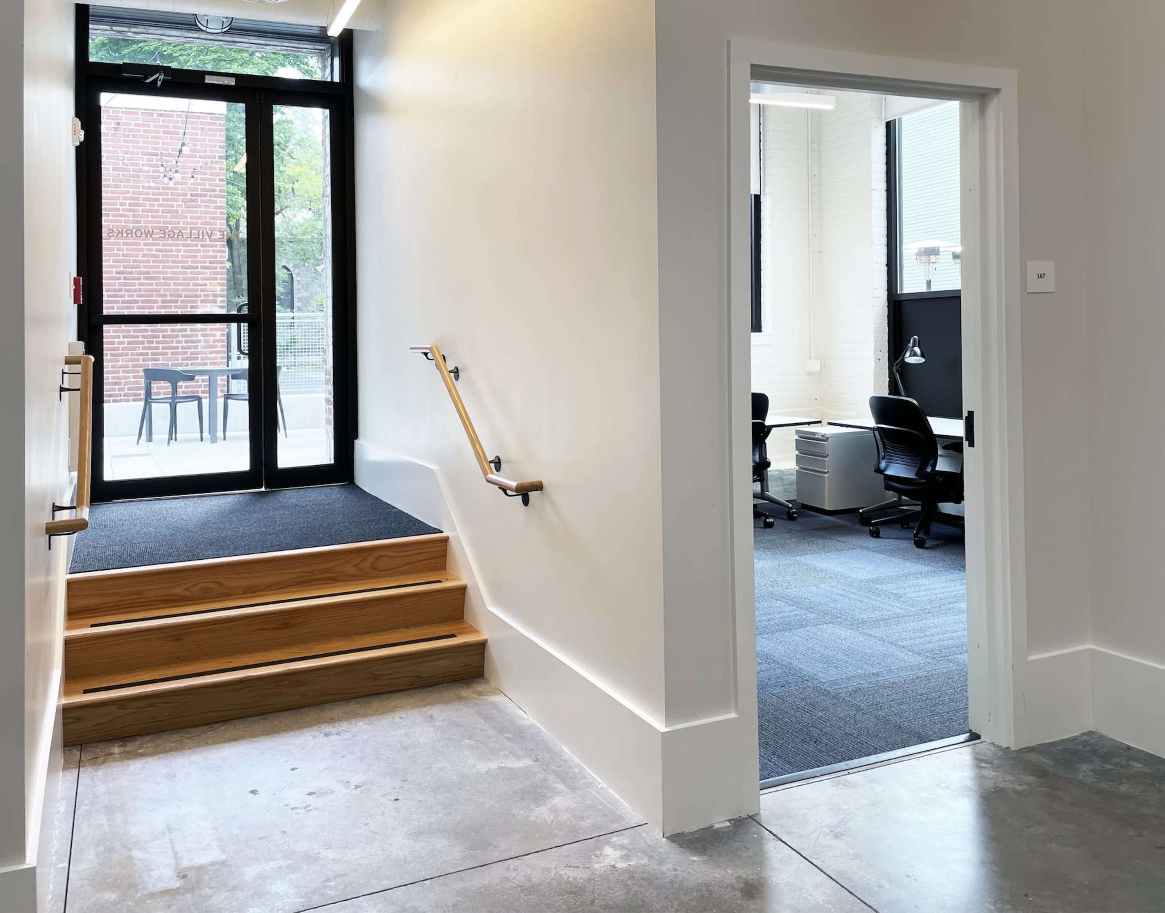 A set of wooden steps leads down from a brightly lit hallway to a door that opens into an office space with chairs and a desk.