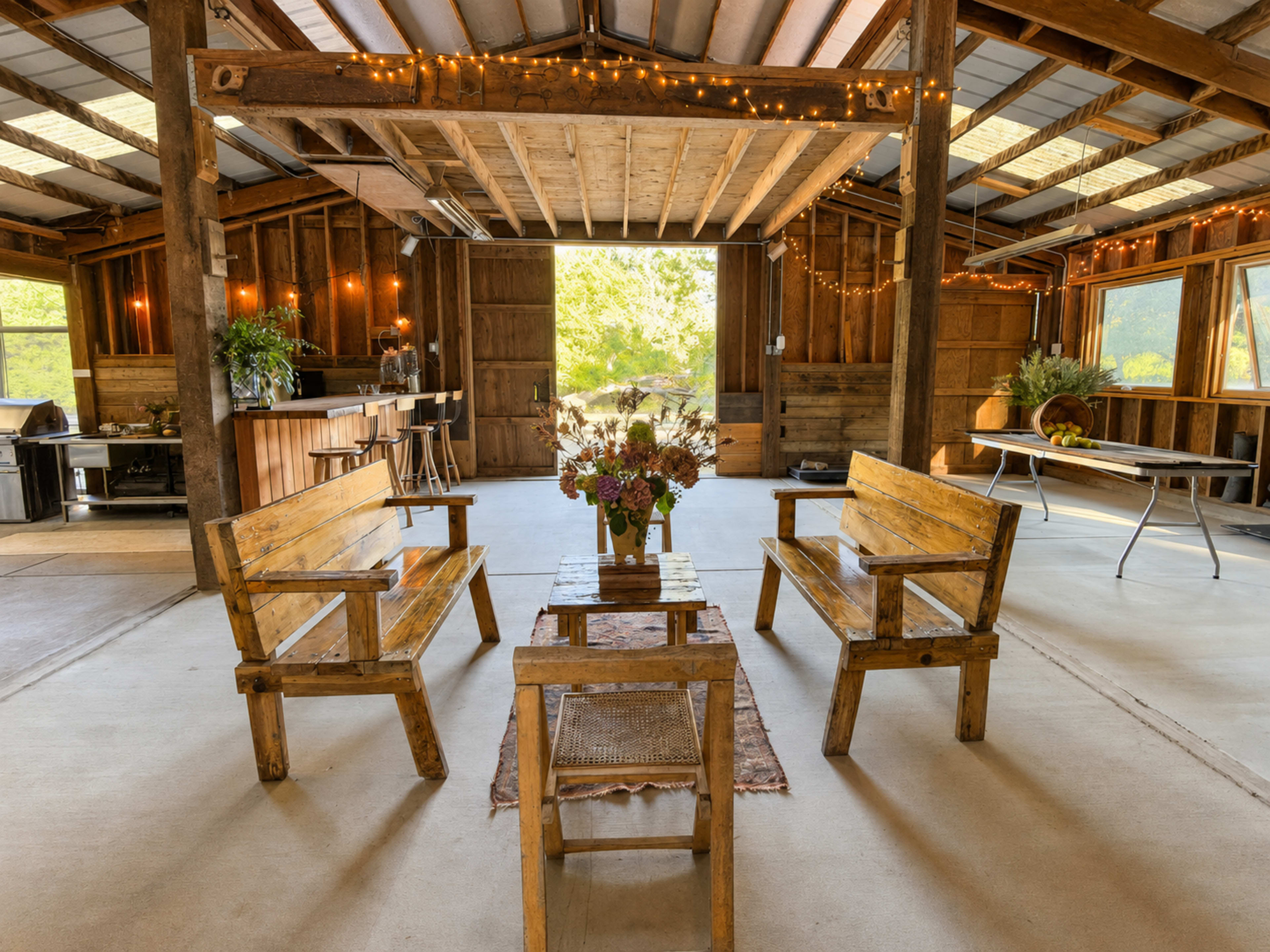 The image shows a rustic interior of a barn-like space with wooden benches arranged around a small table, surrounded by ambient lighting and greenery visible through large windows.