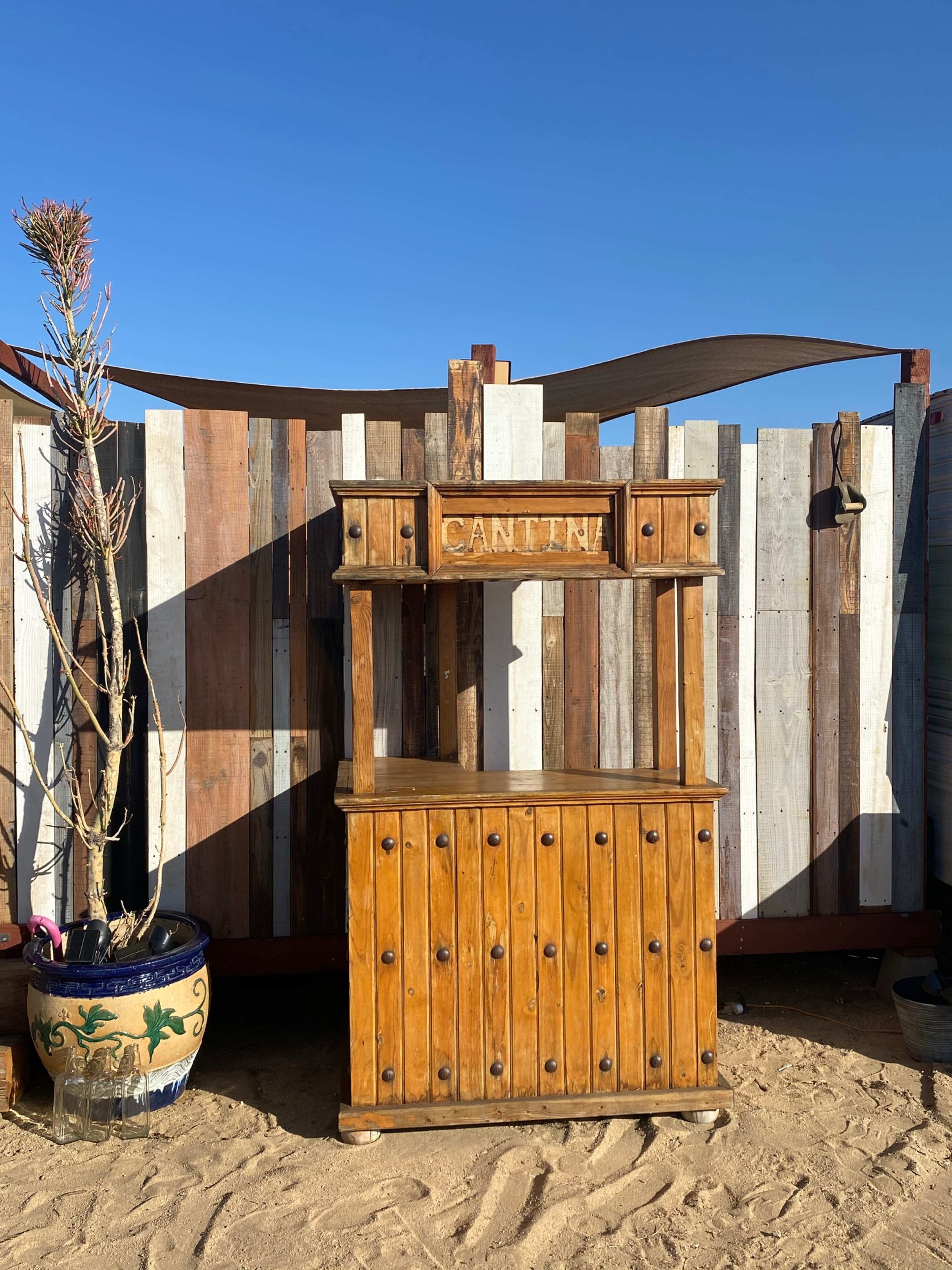 A wooden cantina counter sits against a backdrop of a wooden fence with varying colors, under a clear blue sky.