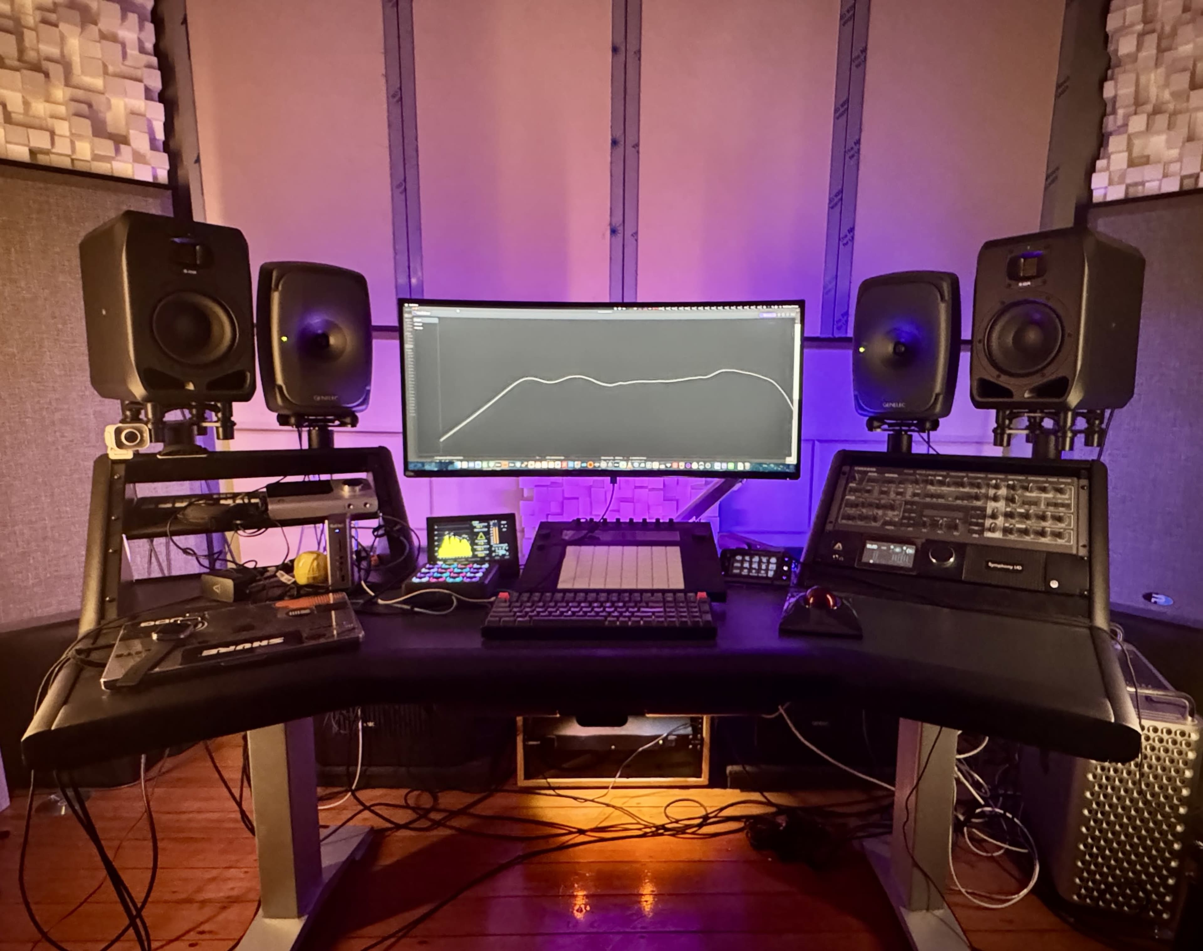 The image shows a modern audio mixing workstation with two studio monitors, a large computer screen displaying audio software, and various audio equipment arranged on a dark desk.