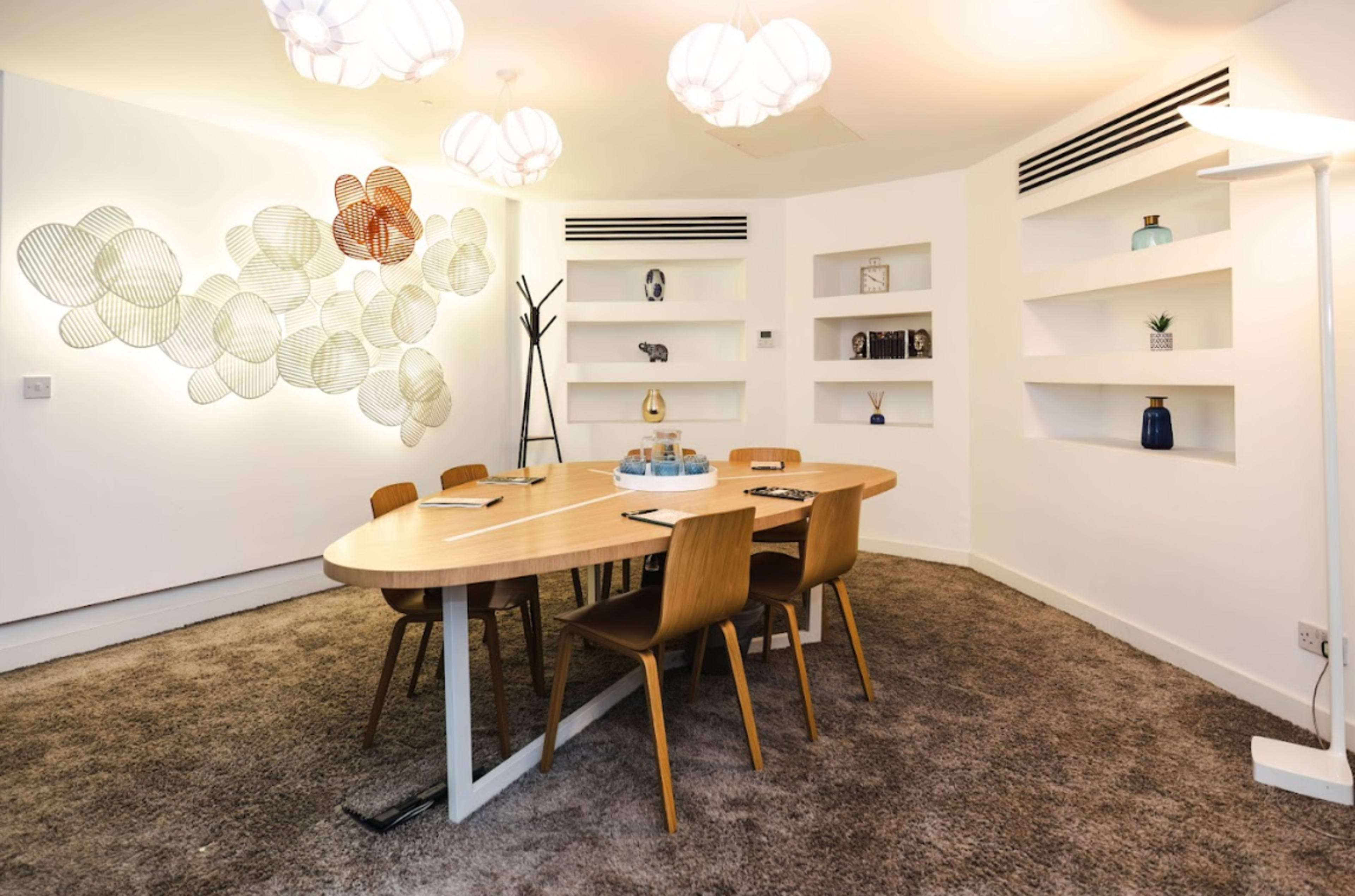 A modern conference room features a round wooden table surrounded by four wooden chairs, with decorative wall art and built-in shelves displaying various objects.
