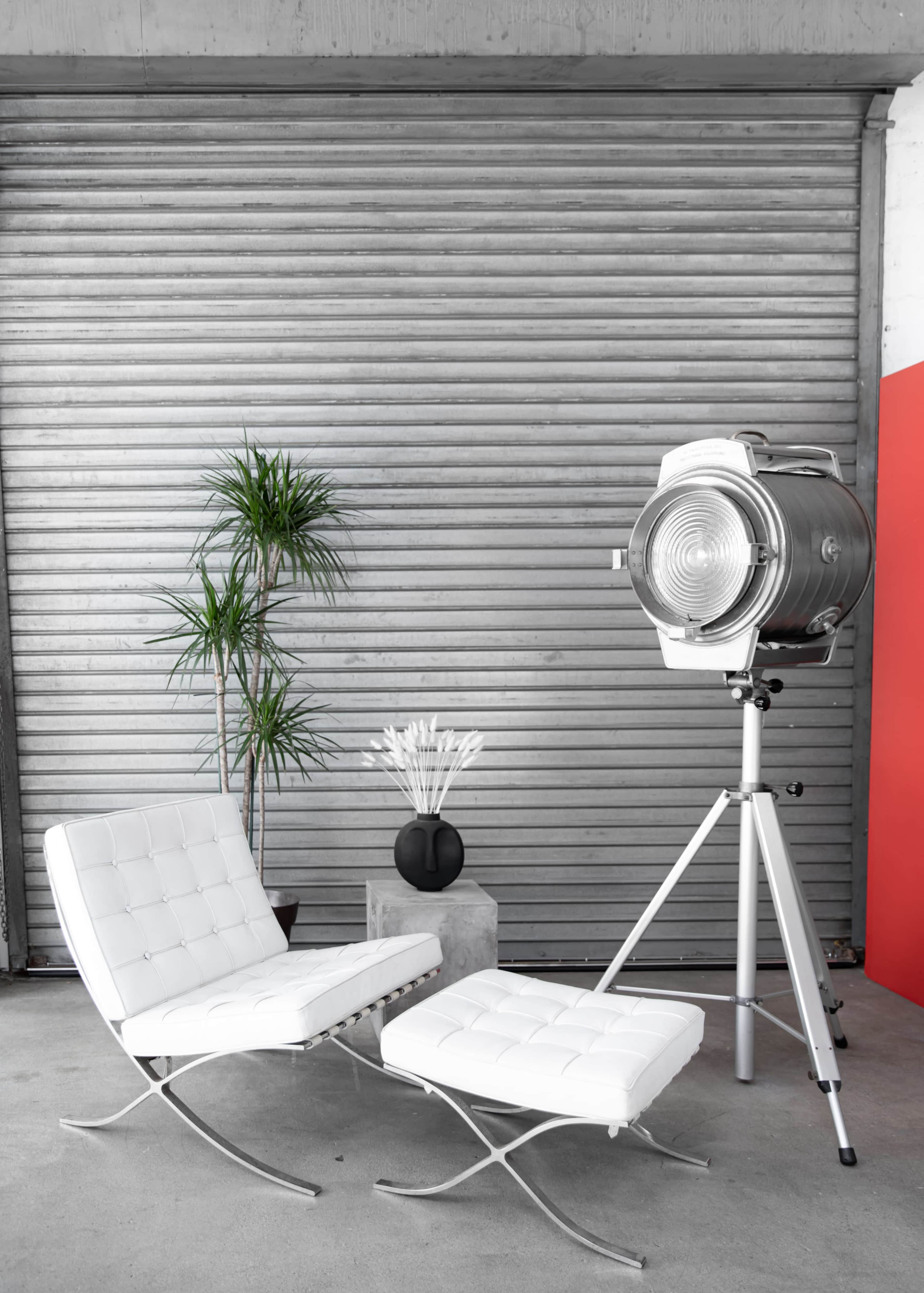 Two modern white chairs beside a large studio light on a tripod, positioned next to a potted plant and a cylindrical black vase against a gray backdrop.
