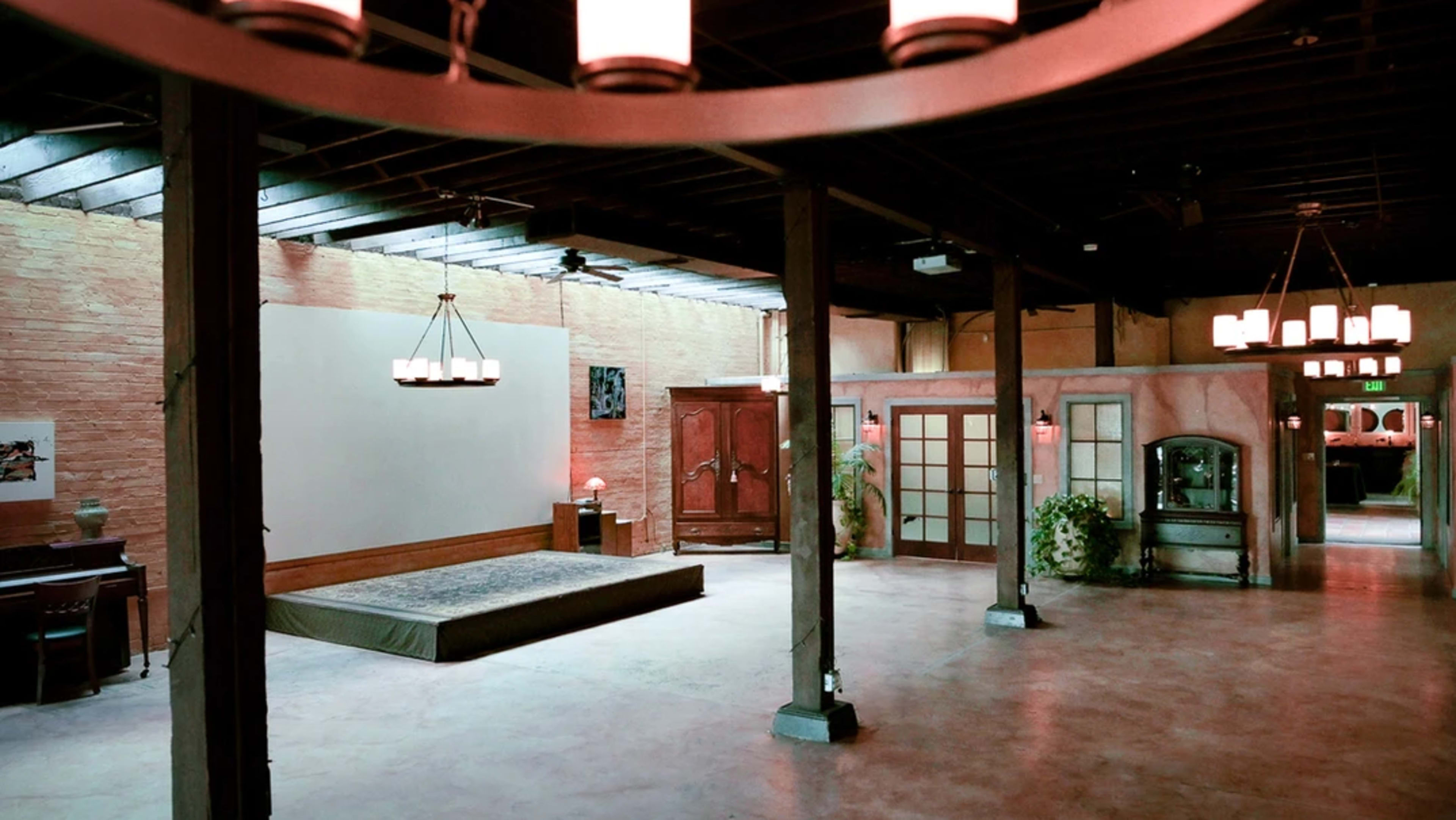 An empty, spacious room with wooden beams, a raised platform at one end, and large glass doors leading to adjacent areas.