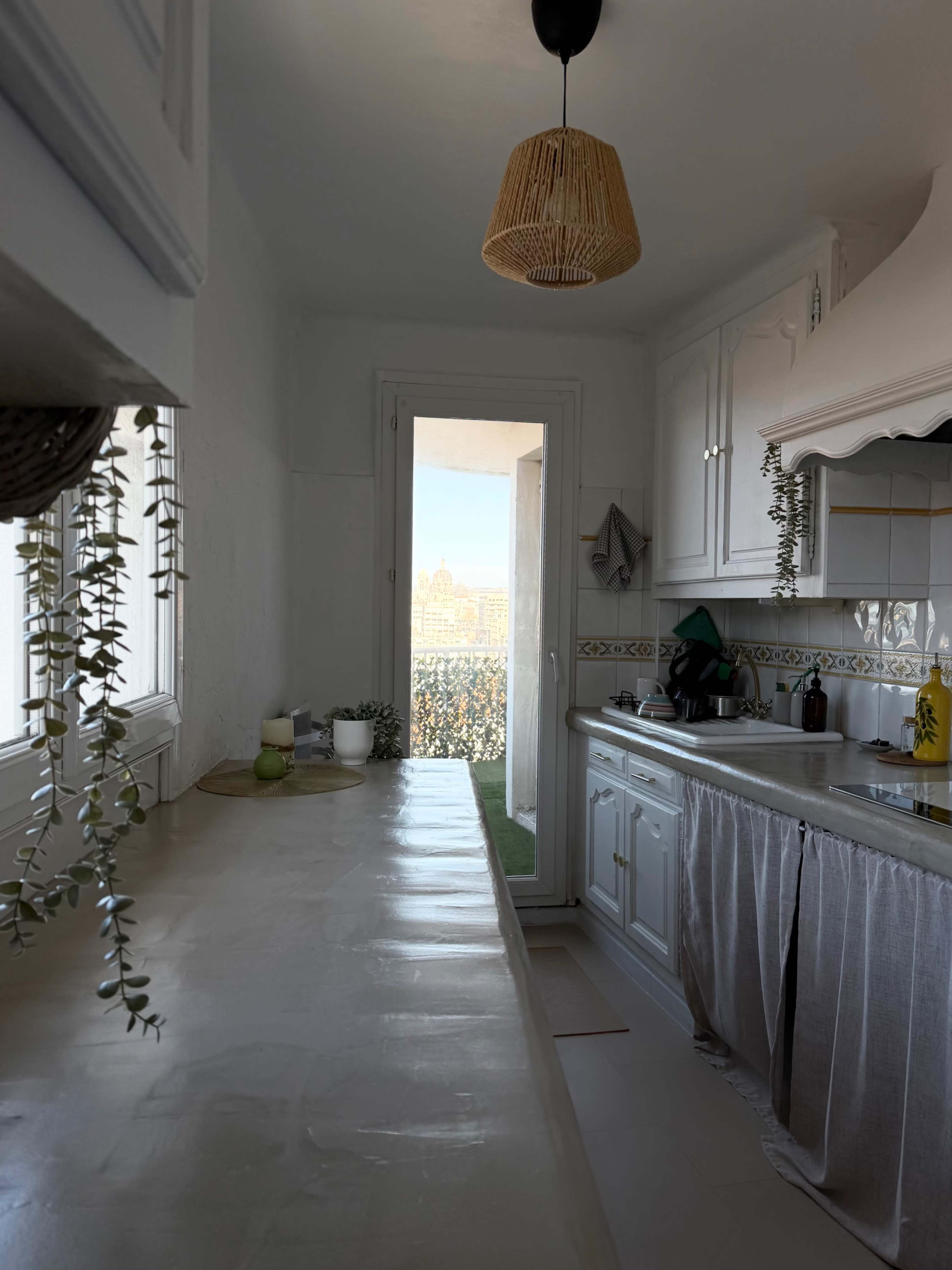 The image shows a bright, minimalistic kitchen with white cabinetry, a long countertop, and a window leading to a view of an outdoor space or cityscape.
