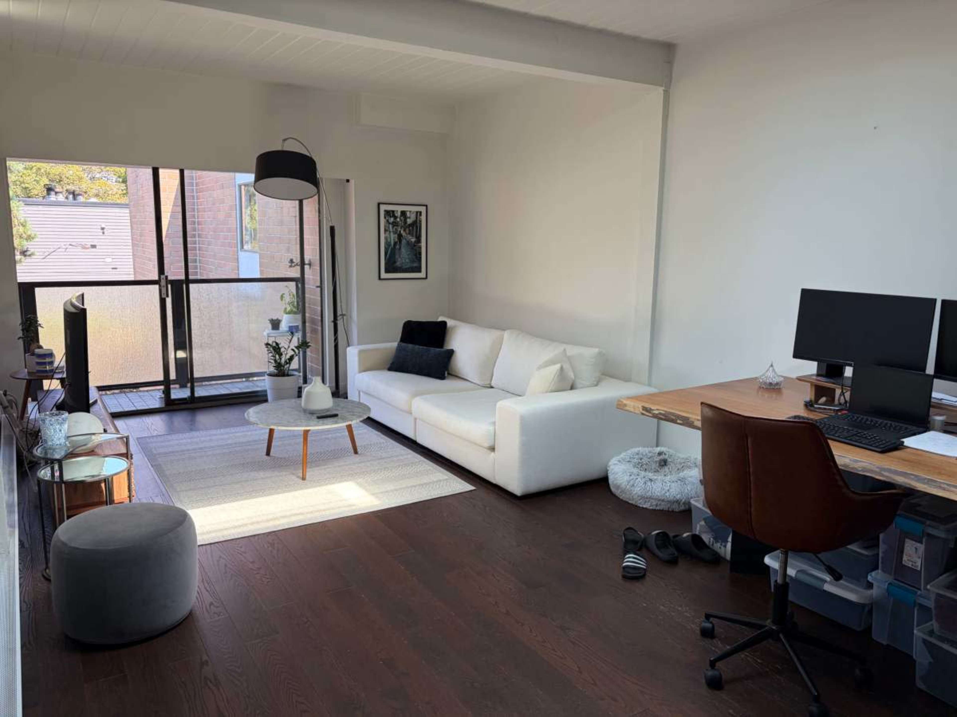 The image depicts a modern living room with a white sofa, a wooden desk with two monitors, and a sliding glass door leading to a balcony.