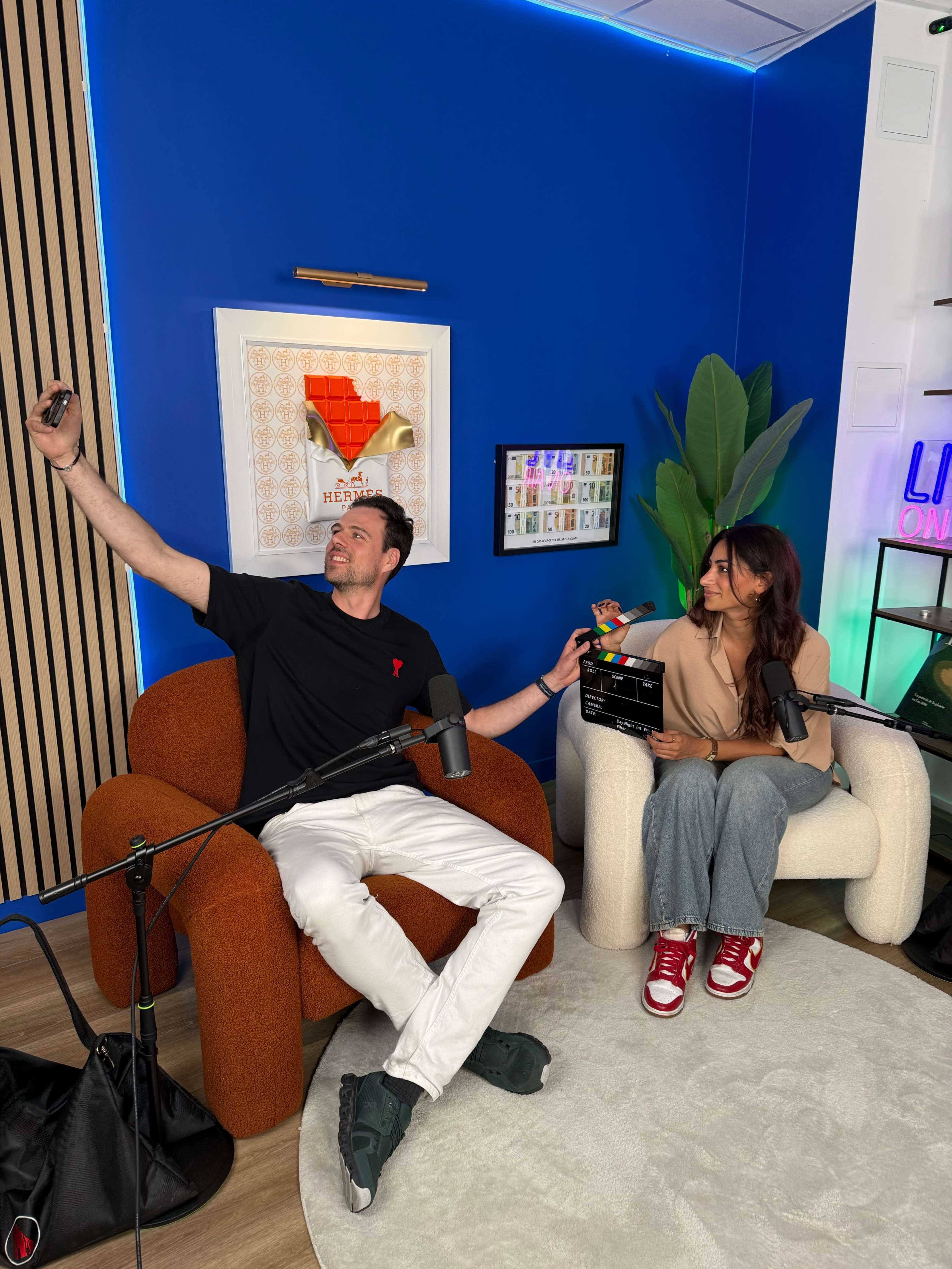 A man and a woman sit in a modern studio, with the man taking a selfie while the woman holds a clapperboard, both surrounded by microphones and decorative elements.
