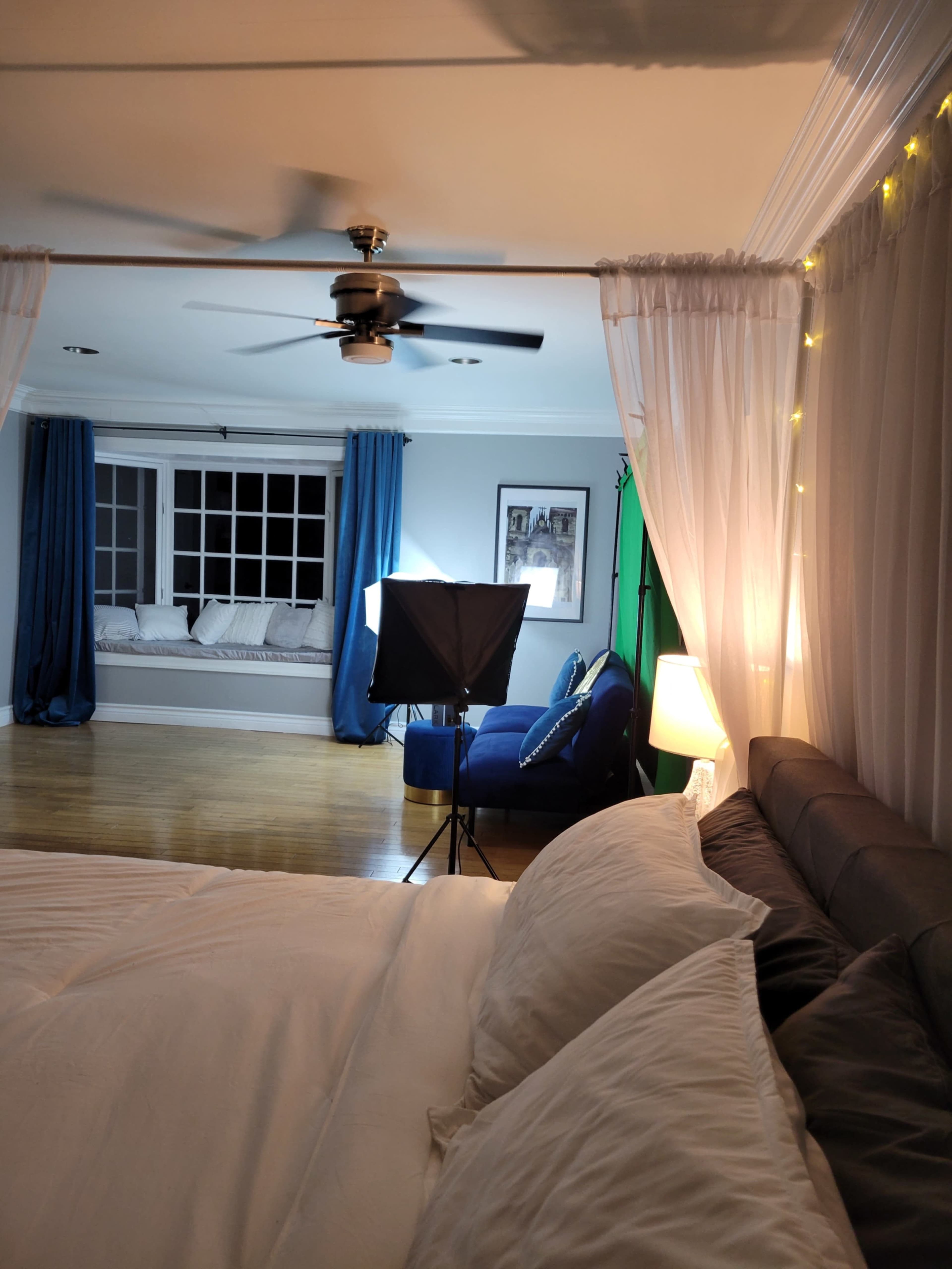 A neatly arranged bedroom features a bed in the foreground, with a cozy seating area and lighting setup visible in the background.