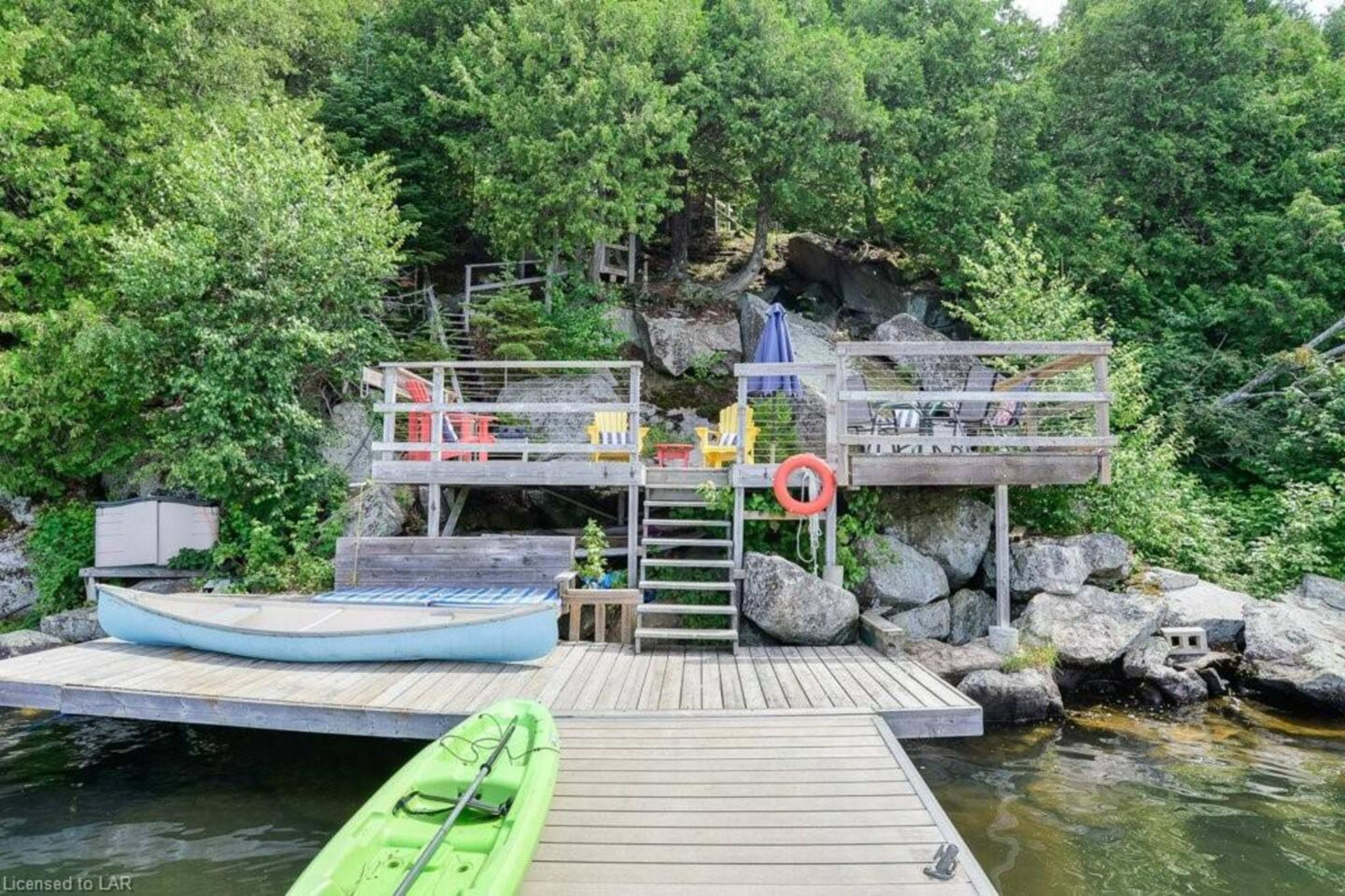 A wooden dock with kayaks, leading to a multi-level deck surrounded by greenery and rocks.