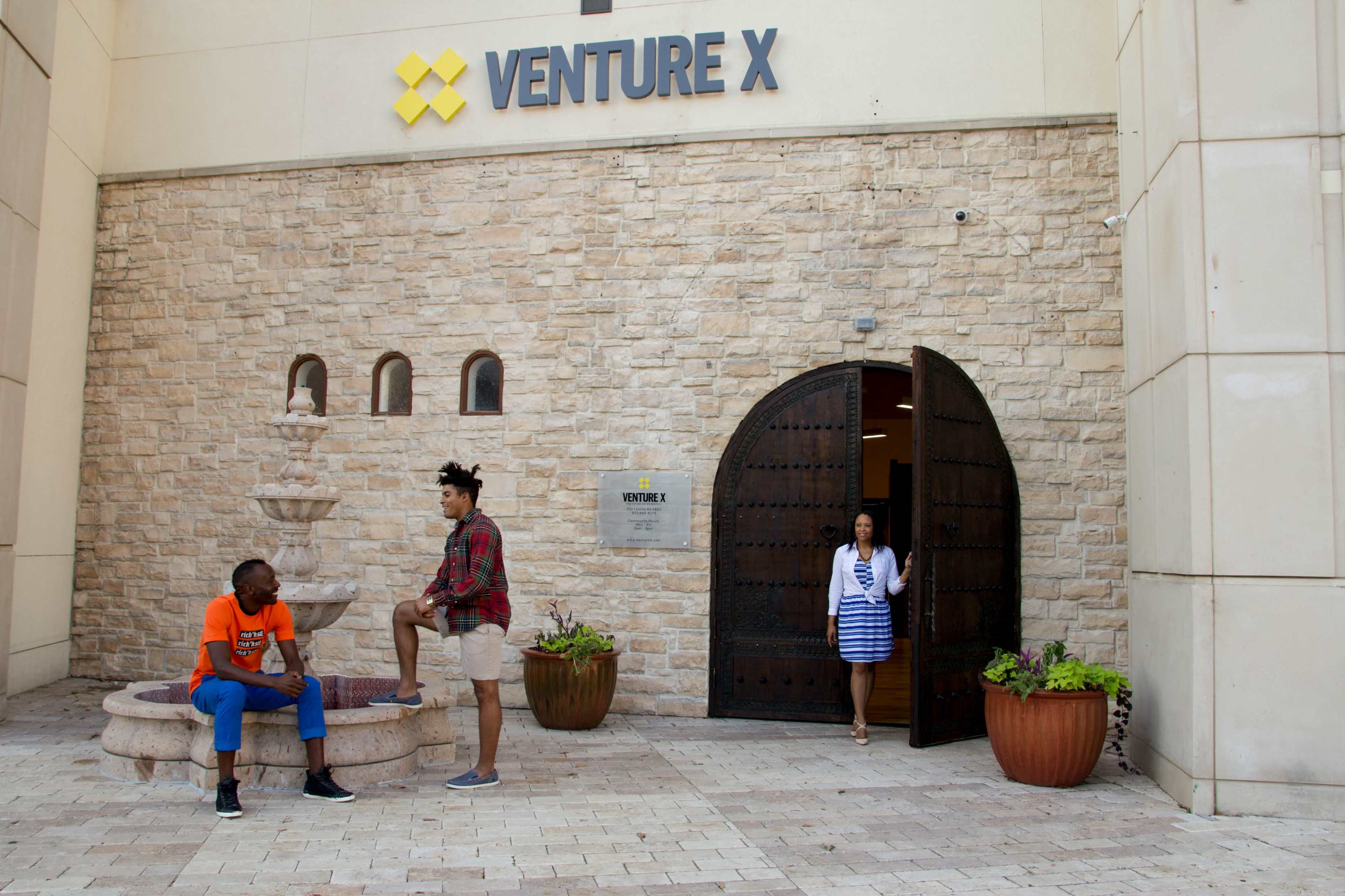 Two men are seated near a fountain outside a building with a sign that reads "VENTURE X," while a woman in a blue and white dress walks through an arched doorway.