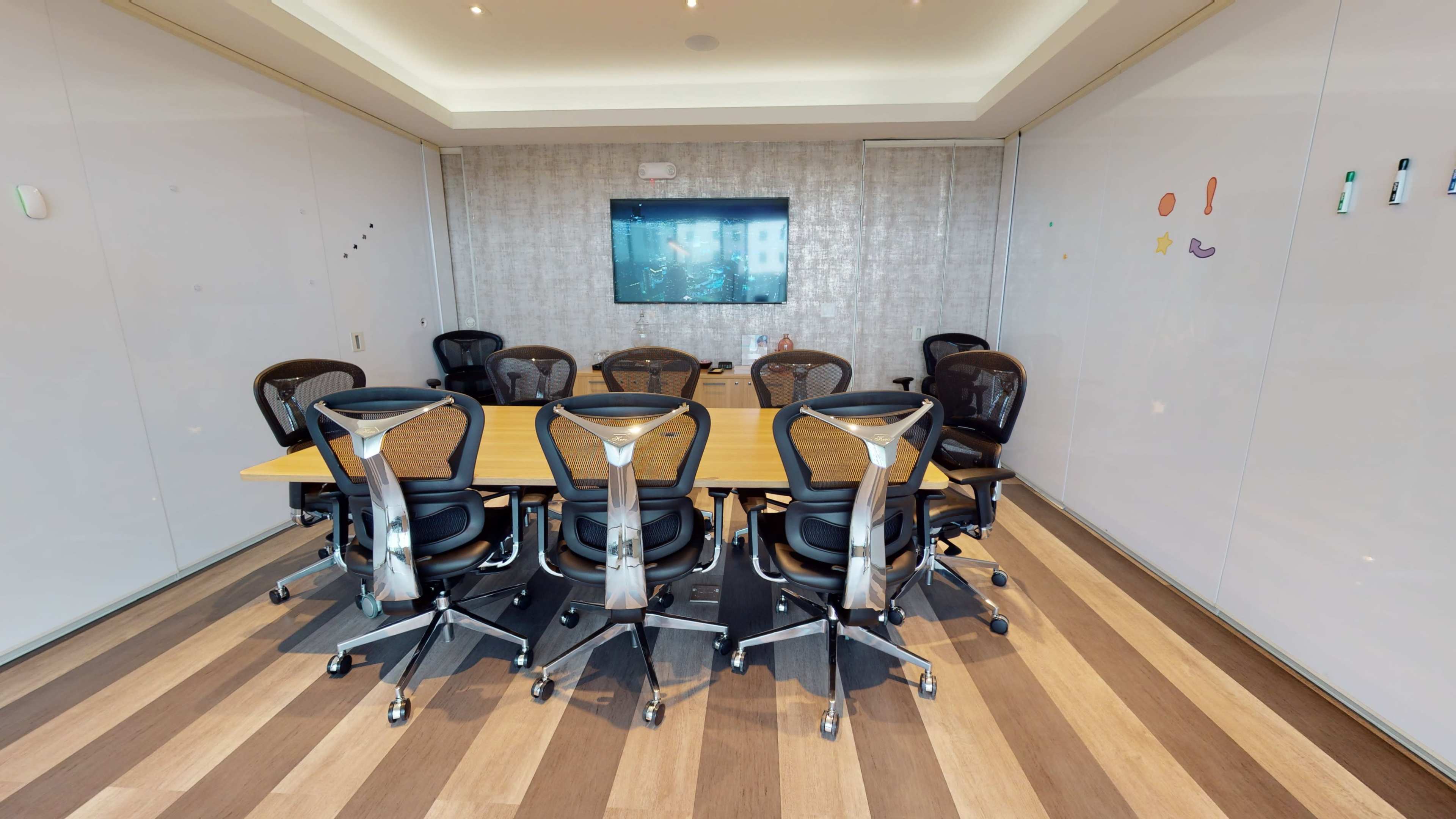 A conference room features a large wooden table surrounded by ergonomic chairs, with a wall-mounted screen in the background.
