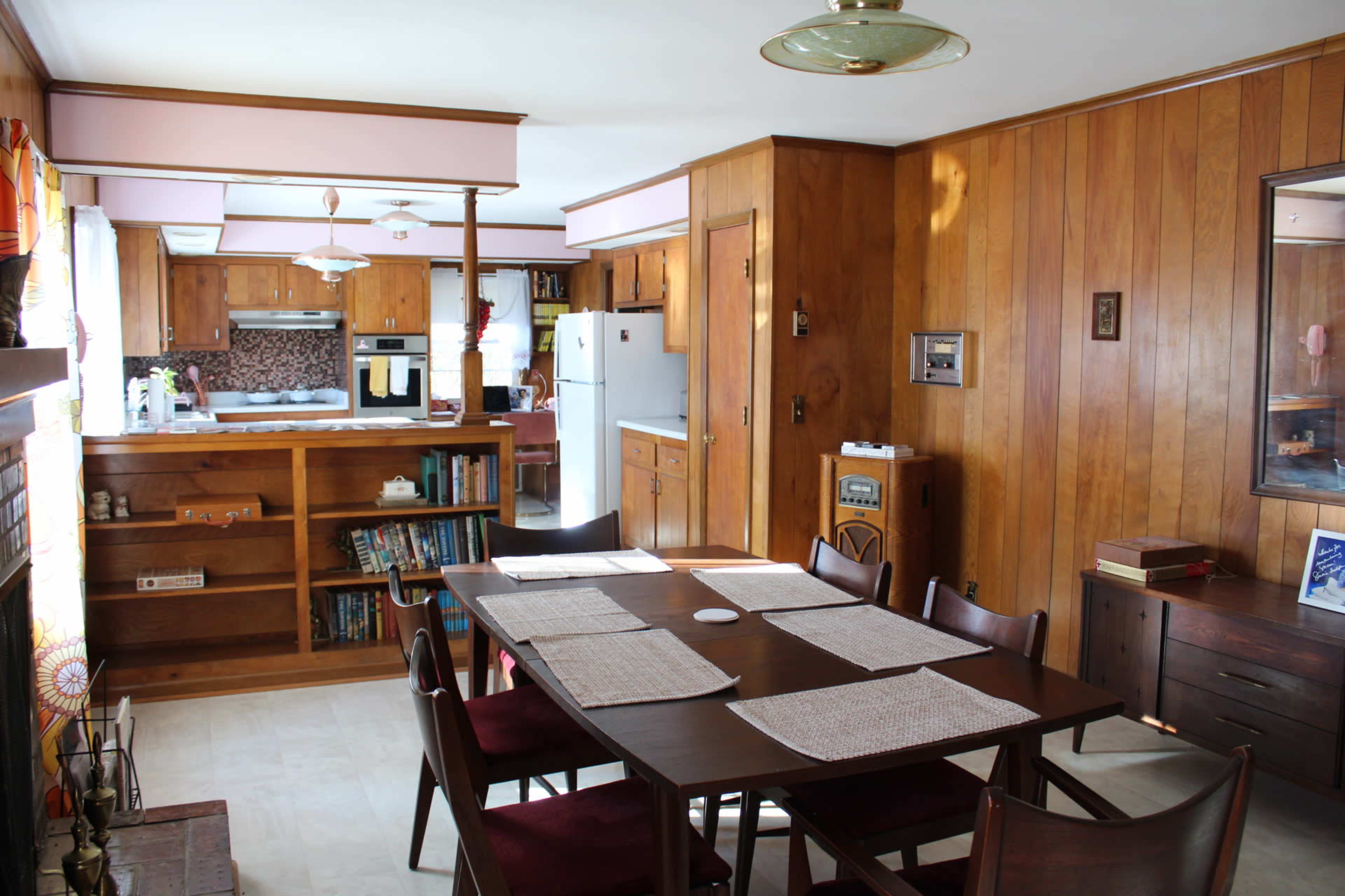 The image shows a wooden-furnished dining area with a table set for meals, adjacent to an open kitchen in a well-lit room.