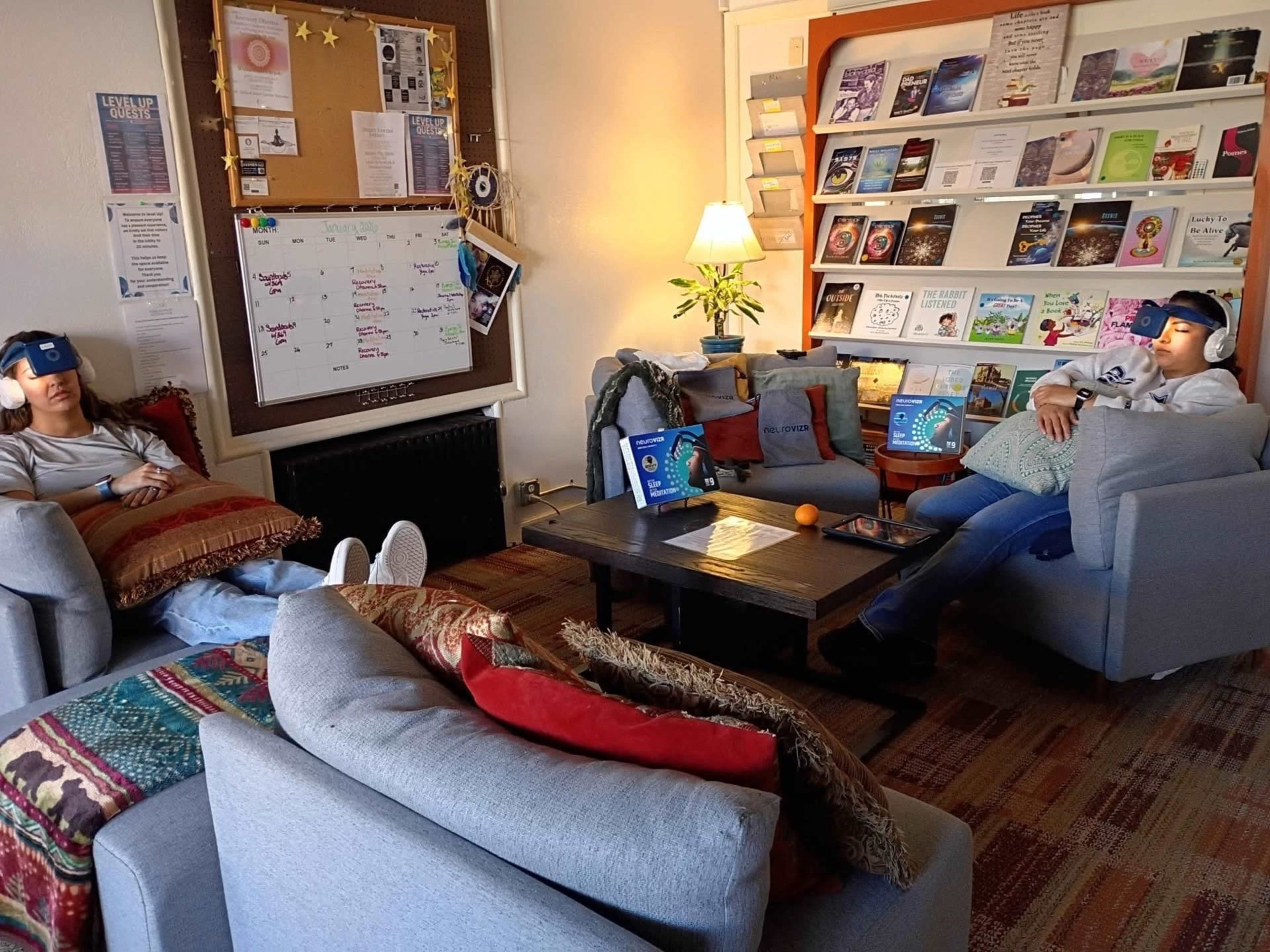 Two people are sitting in a cozy room, wearing headphones and relaxing on comfortable sofas surrounded by bookshelves filled with various books.