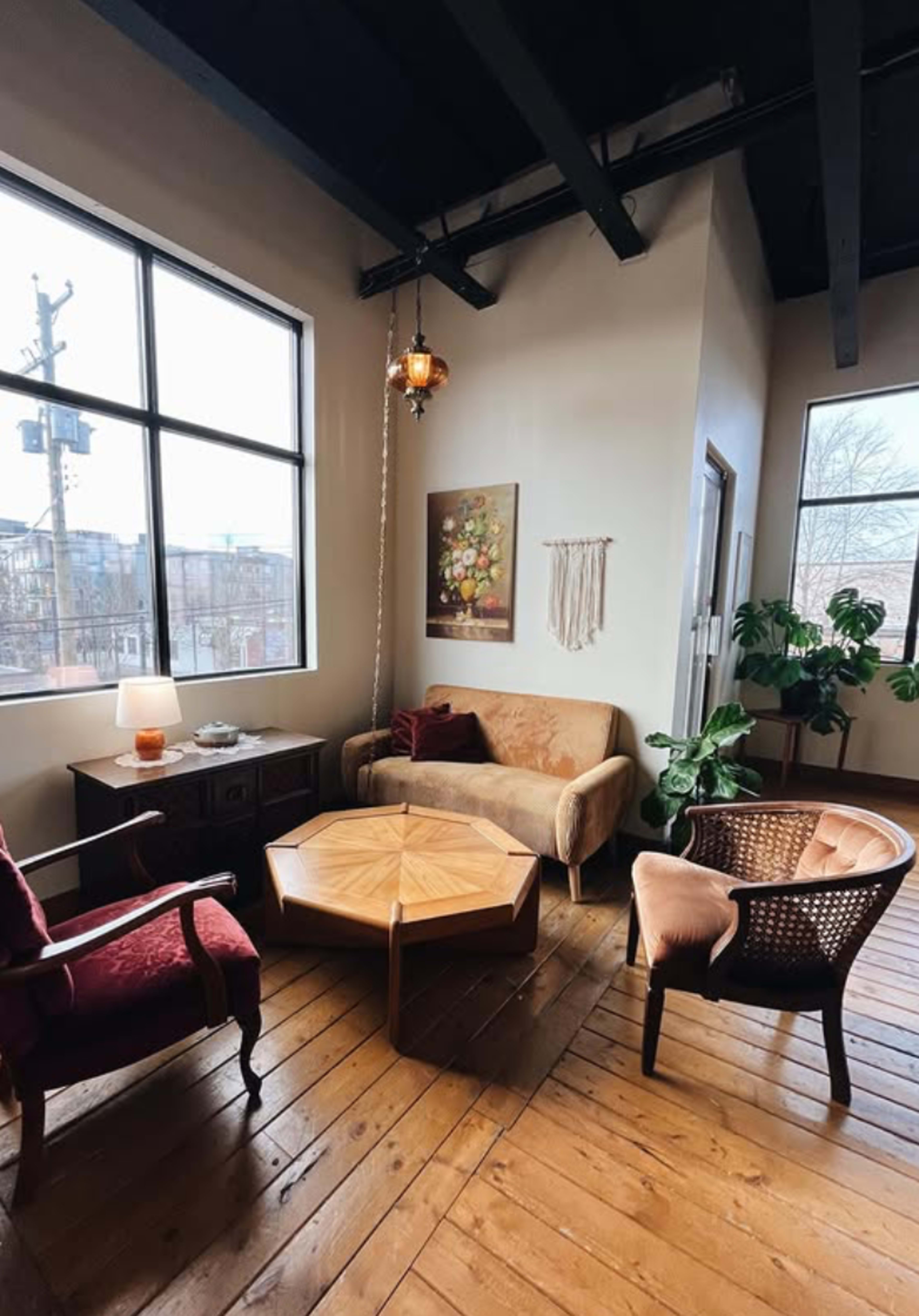 The image shows a cozy living room with a mix of furniture, including a brown sofa, two chairs, a wooden coffee table, and large windows allowing natural light to enter.