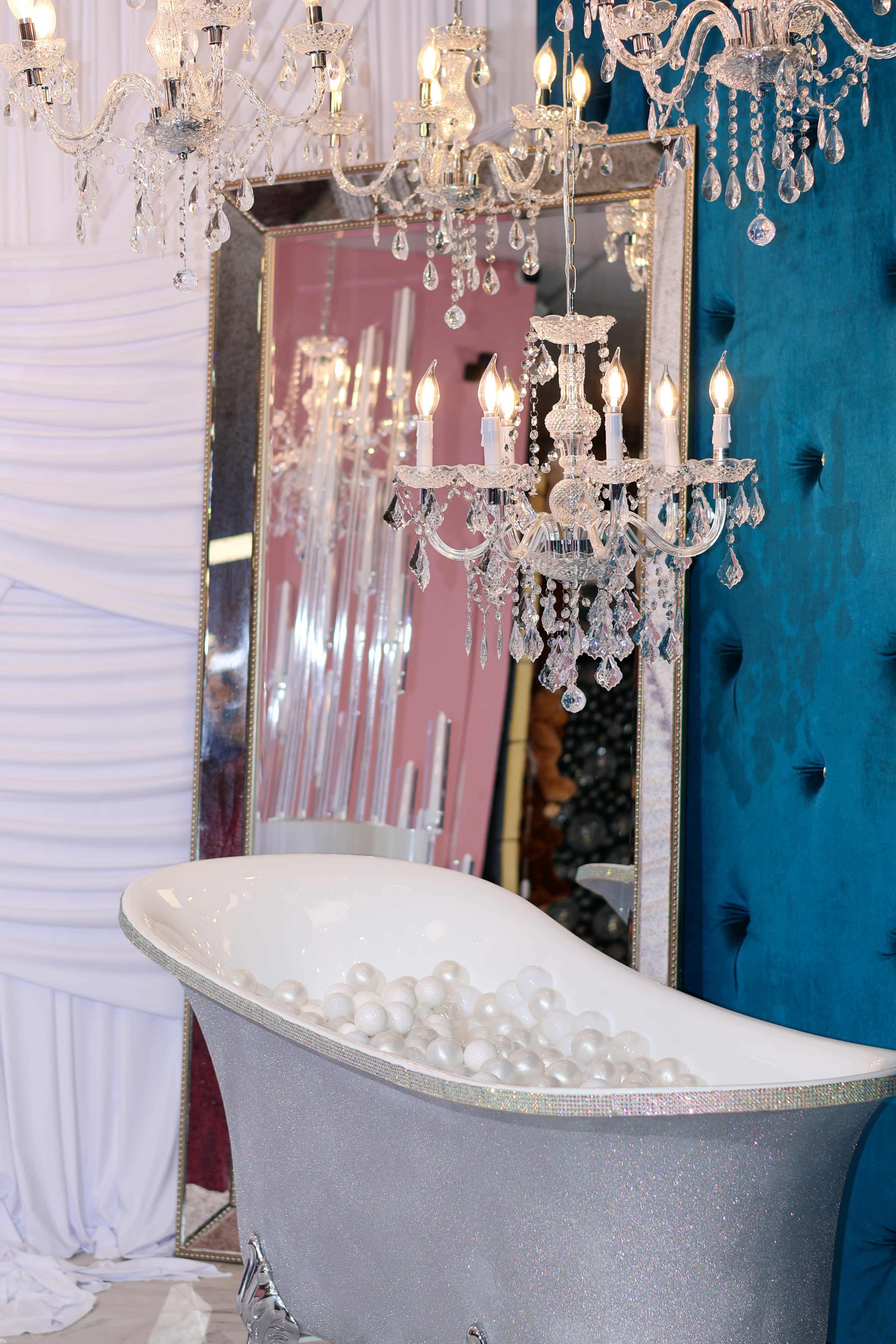 The image shows a stylish bathtub filled with white balls, set against a backdrop of a large mirror and ornate chandeliers.