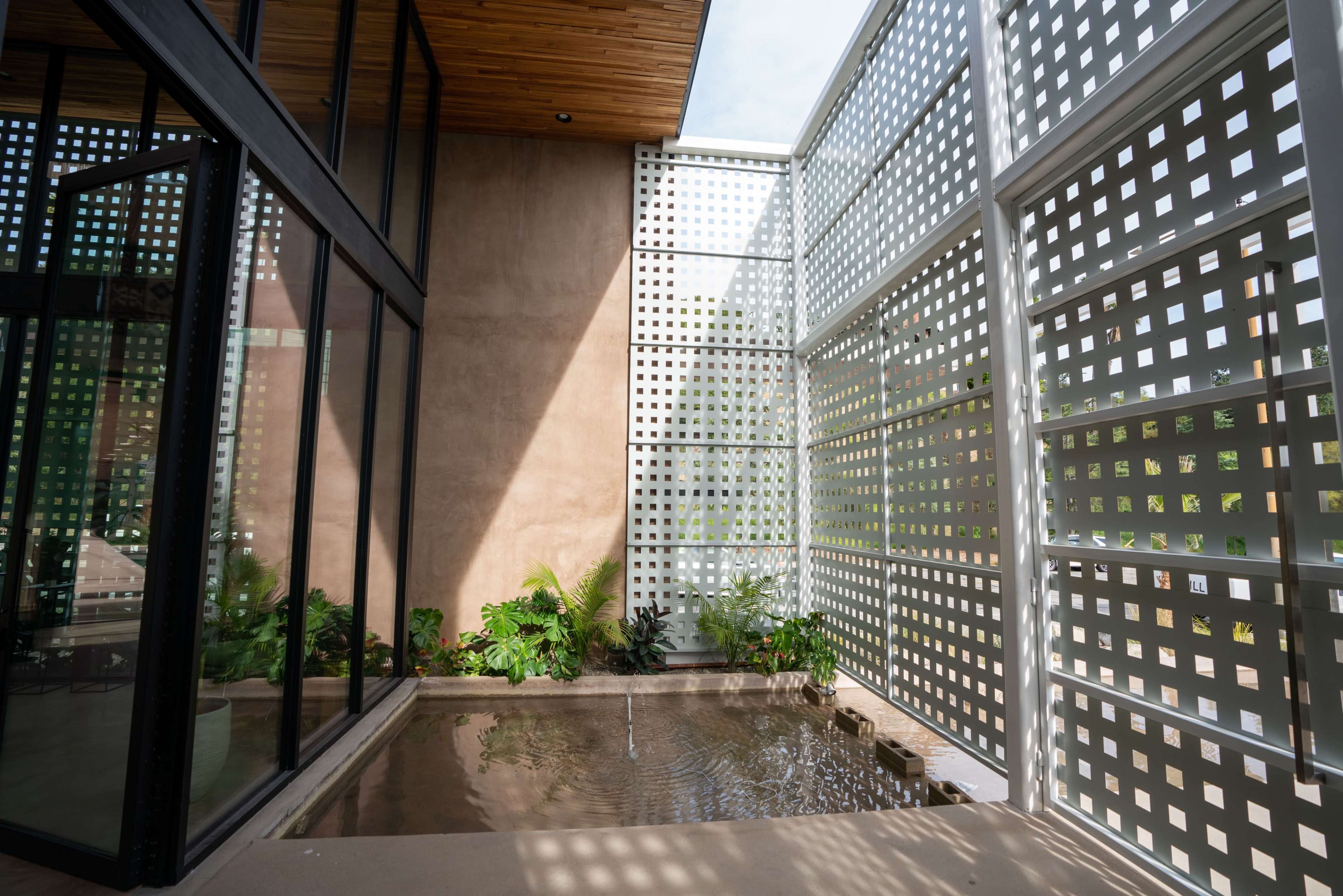 The image shows a modern architectural space with large glass walls, an open water feature, and greenery enclosed by patterned screens.