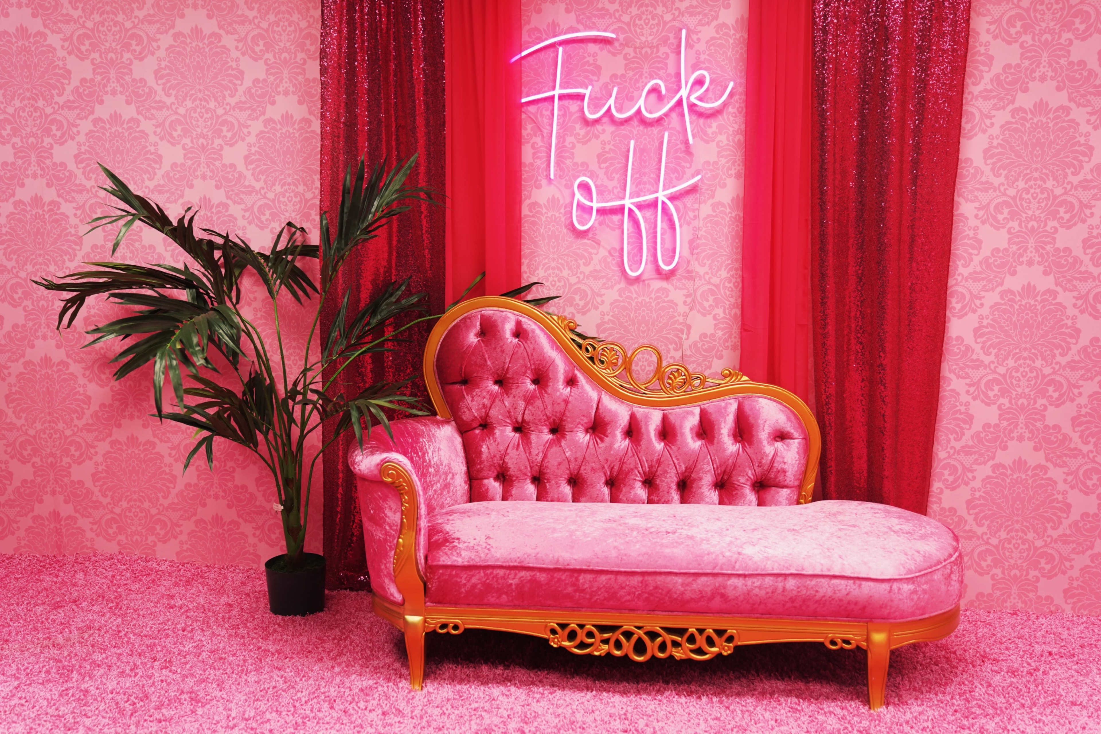 A pink vintage chaise lounge with an ornate frame is placed against a patterned wall, complemented by a neon sign that reads "Fuck off" and a potted plant nearby.
