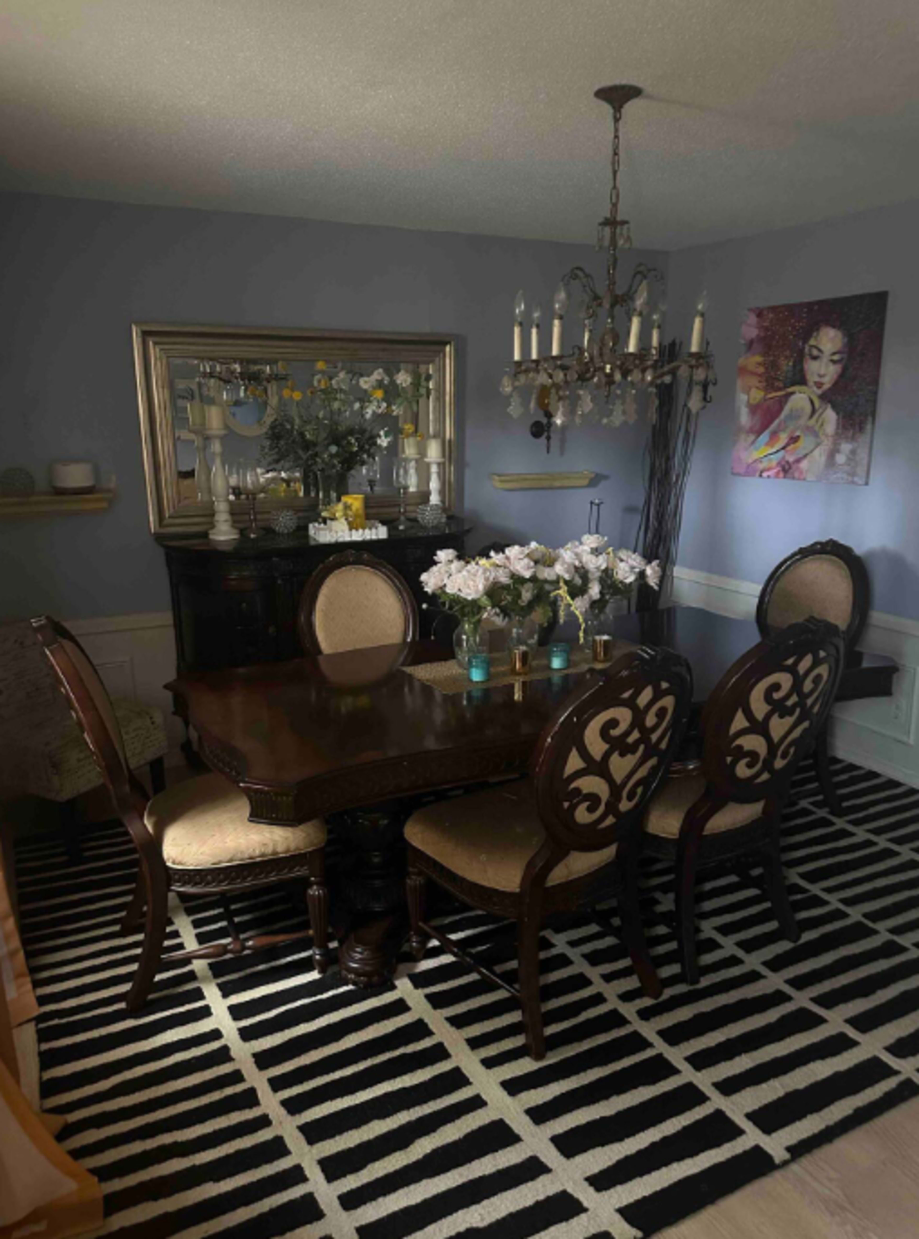 A dining room features a wooden table surrounded by upholstered chairs, with a chandelier overhead and floral arrangements on the table.