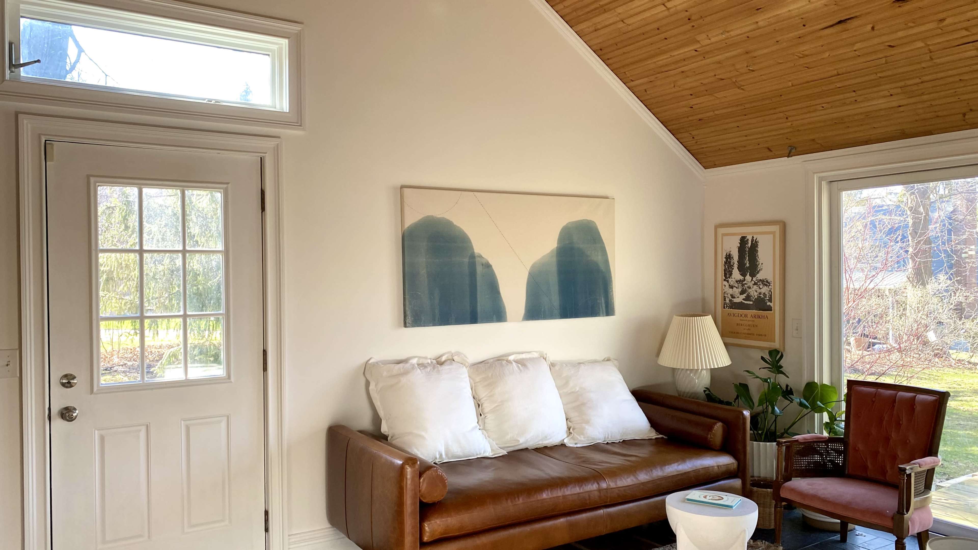 A cozy living room featuring a brown leather sofa with white cushions, a vintage armchair, a table lamp, and a large piece of artwork depicting blue mountains on the wall.