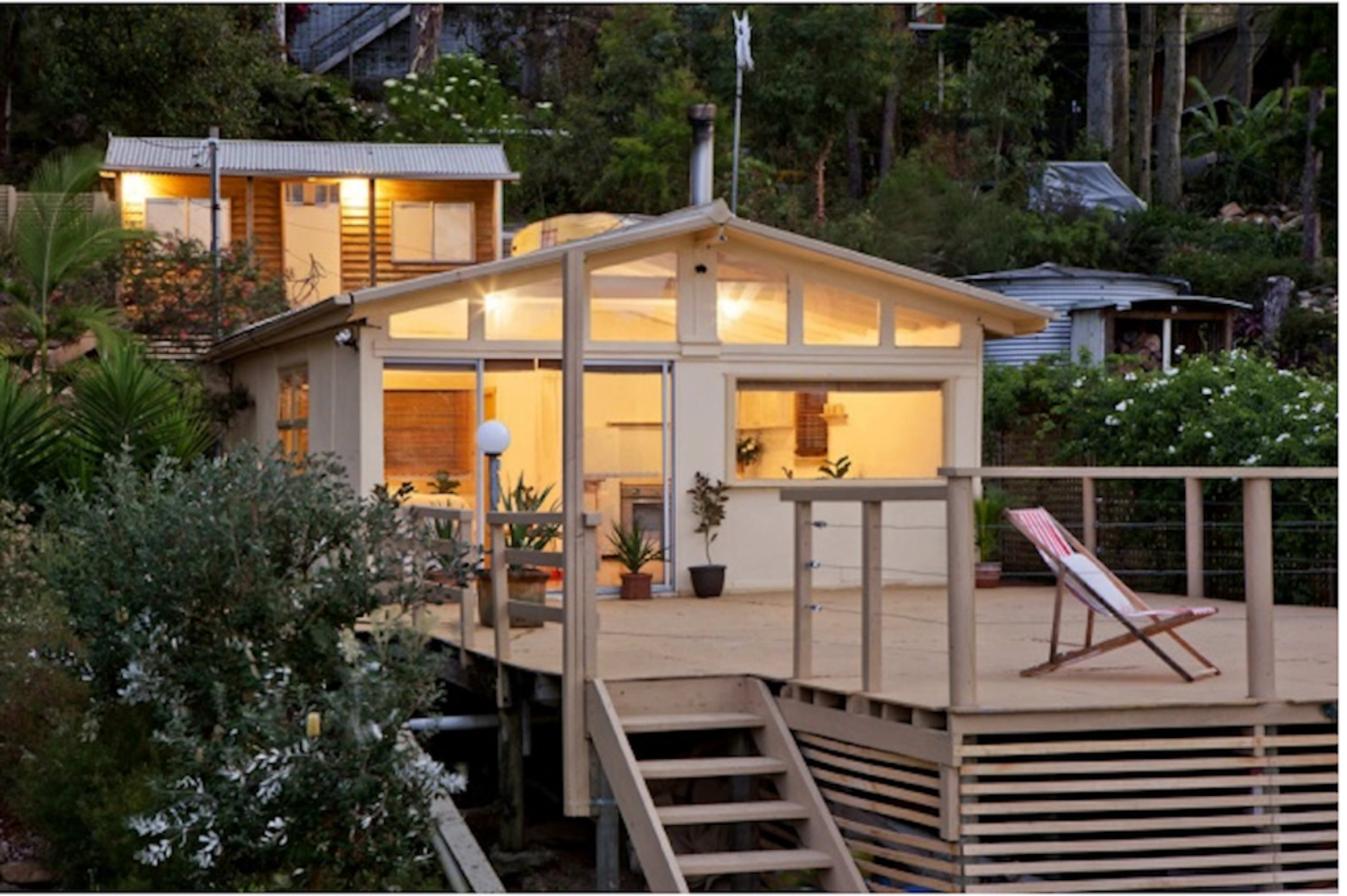A wooden house with a large deck is situated on a hillside, showcasing large windows and surrounded by greenery.