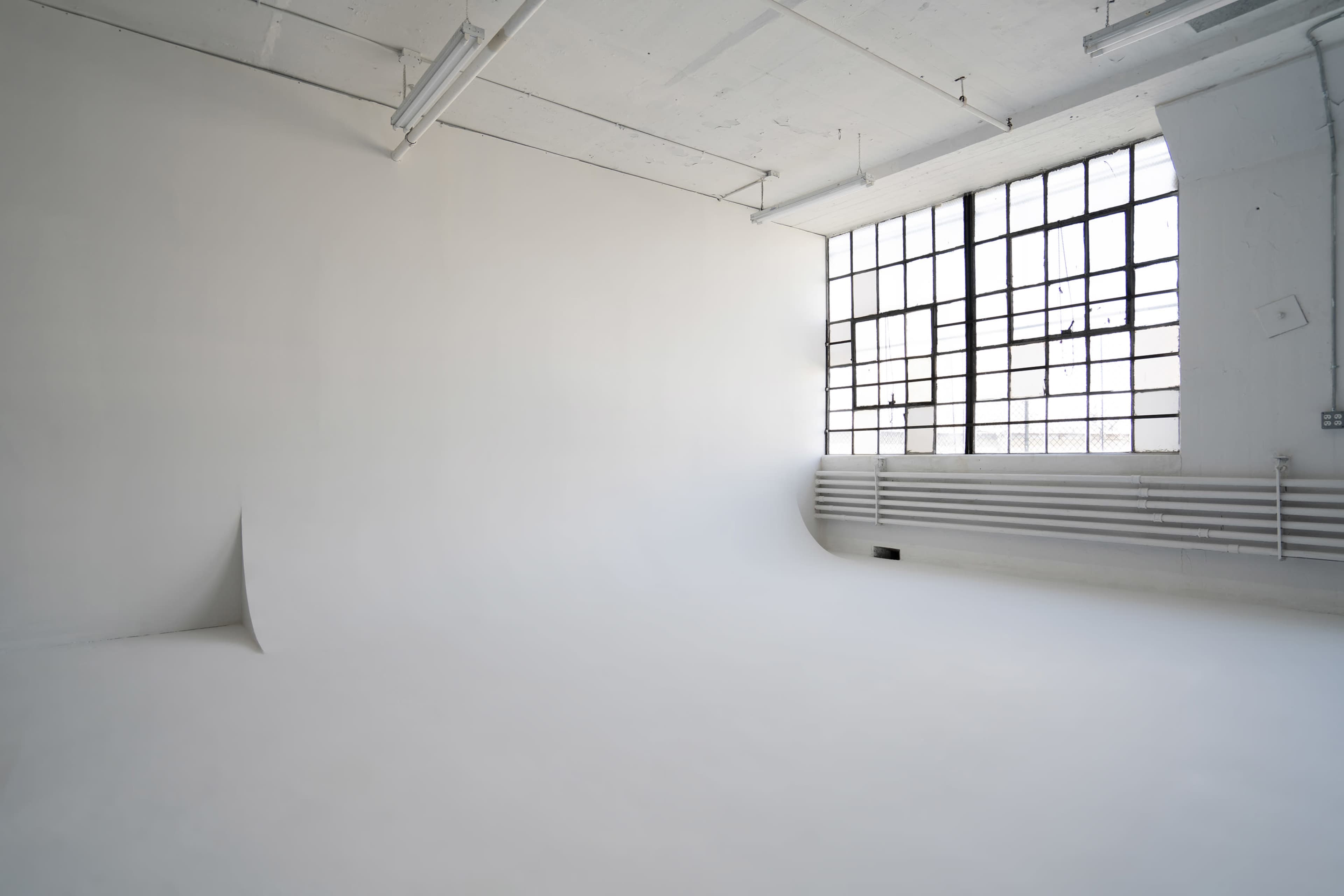A spacious, empty studio features a seamless white backdrop and large industrial windows letting in natural light.